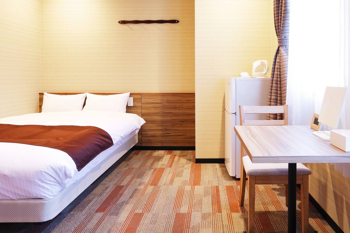 Een hotelkamer met een bed, een bureau, een stoel en een klein tafeltje met een lamp en een doos.
