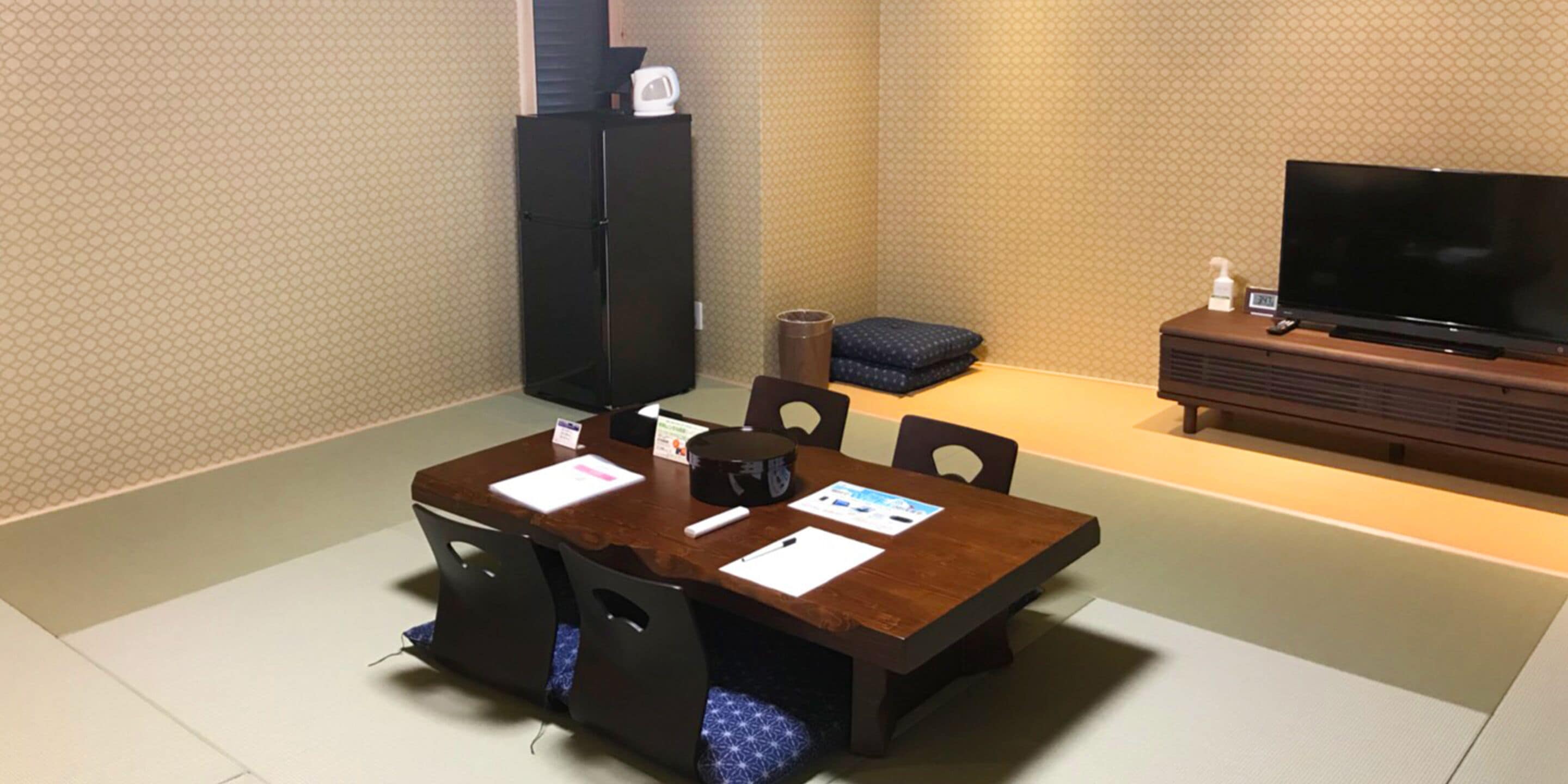 A hotel room with a wooden dining table, chairs, a black refrigerator, a television, and a small blue sofa.