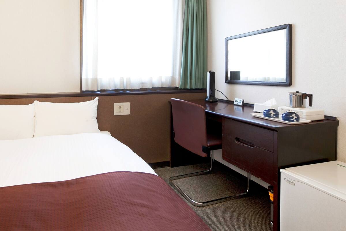 Hotelkamer met een bed, bureau, stoel, kleine koelkast, spiegel en raam met gordijnen.