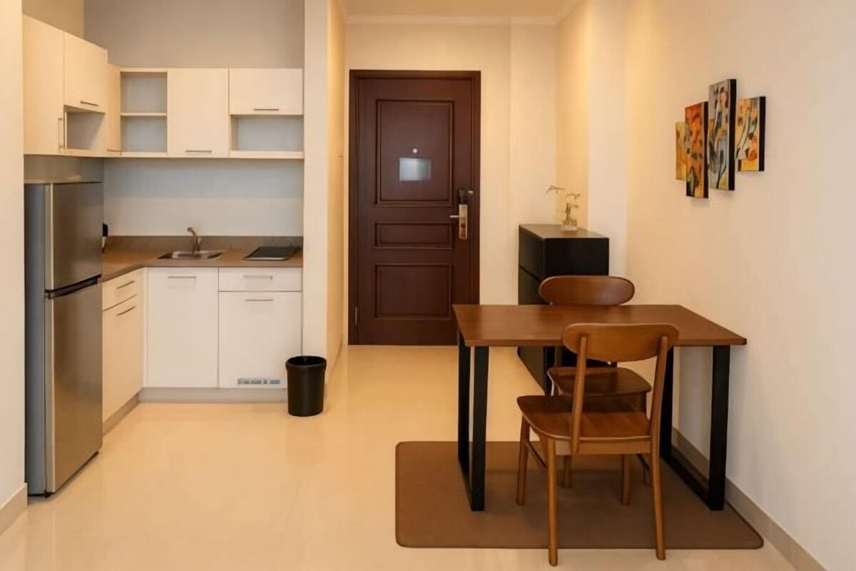 Cocina compacta con refrigerador, estufa, pileta y muebles de cocina blancos. Hay una mesa de comedor con sillas y una puerta que da a otra habitación.