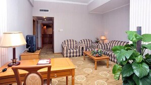 Habitación de hotel con un sofá de estampado de cuadros, un escritorio de madera con lámpara, una mesita con un arreglo floral y una planta verde grande.