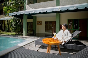 A person sitting on a lounge chair by a poolside with a table and a drink. - Spazzio Bali Hotel (Legian)