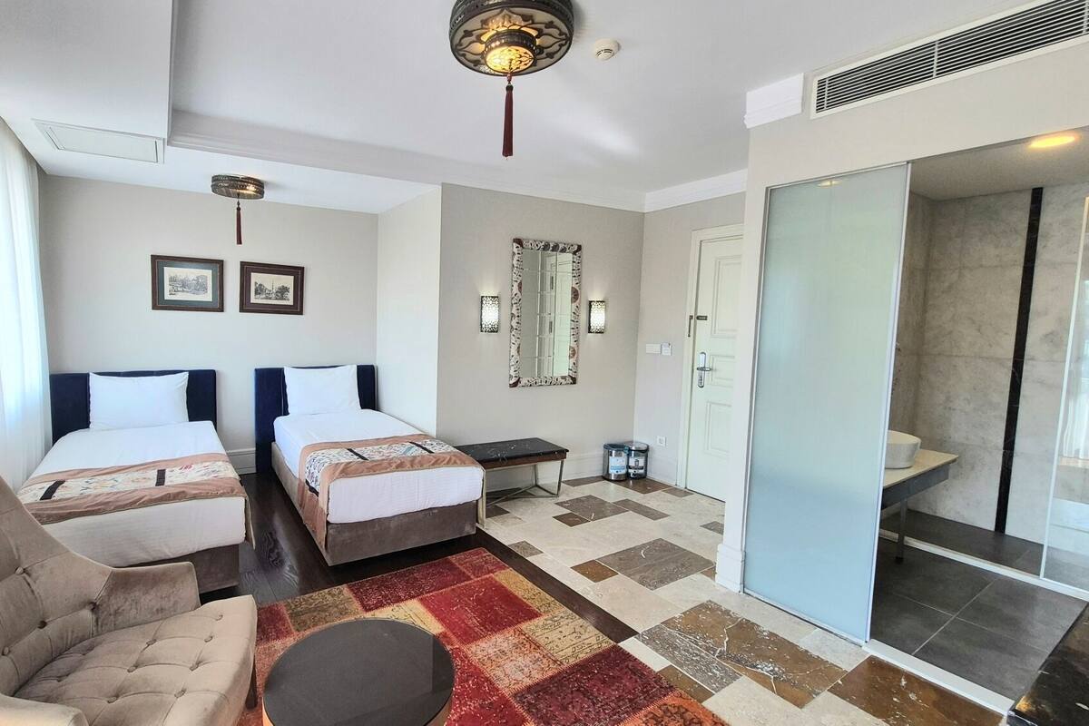A hotel room with two beds, a chair, a small table, a mirror, and a bathroom with a glass door.