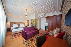 A hotel room with a bed, a purple sofa, a wooden dining table, a small round table, a brick wall, and a ceiling with decorative molding. - Azade Suites (Istanbul)