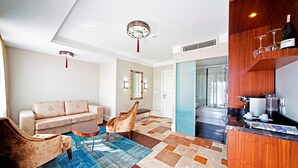 Un salon avec un canapé, un fauteuil et une table basse, un coin cuisine doté d’un plan de travail en bois et de verres à vin, et une salle de bain visible à travers une porte ouverte.