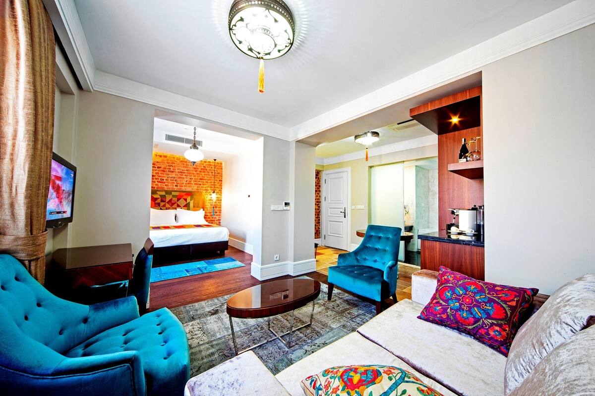 A modern hotel room with a sofa, armchair, and a small table. There is a TV mounted on the wall, a brick wall in the background, and a door leading to another room.
