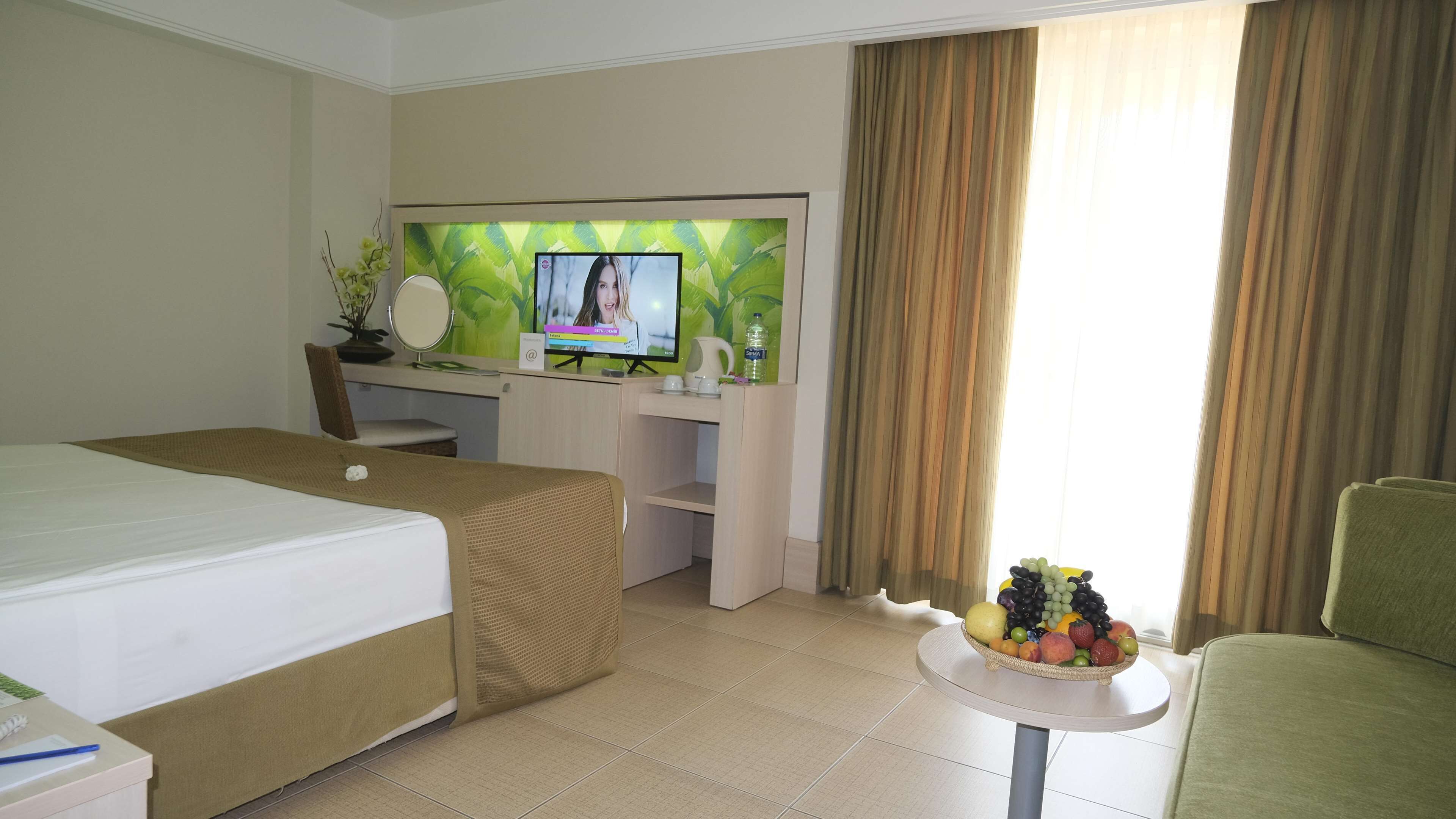 A hotel room with a bed, a desk with a computer, a chair, a small table with fruit, and a window with curtains.