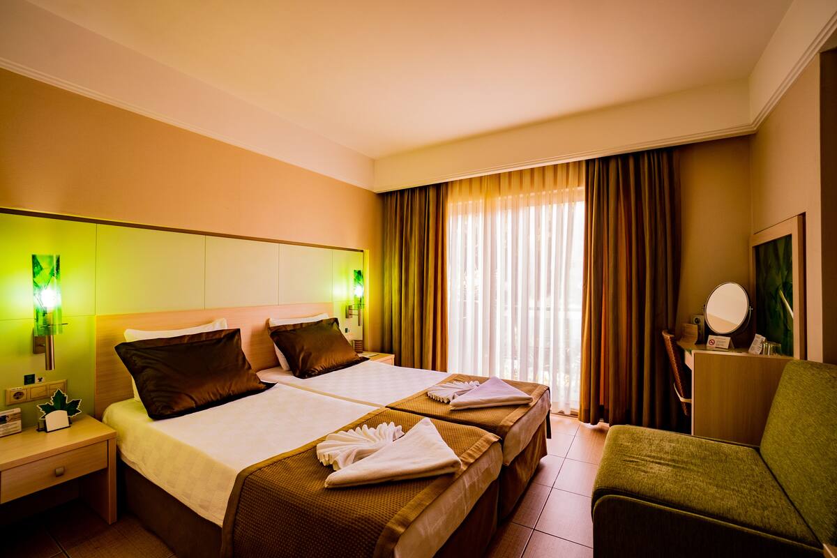 A hotel room with two beds, a green armchair, a desk with a mirror, and a window with curtains.