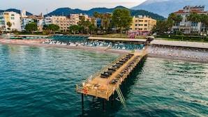 A wooden pier extending into a body of water with rows of chairs and tables, surrounded by buildings and palm trees. - Ambassador Plaza (Kemer)