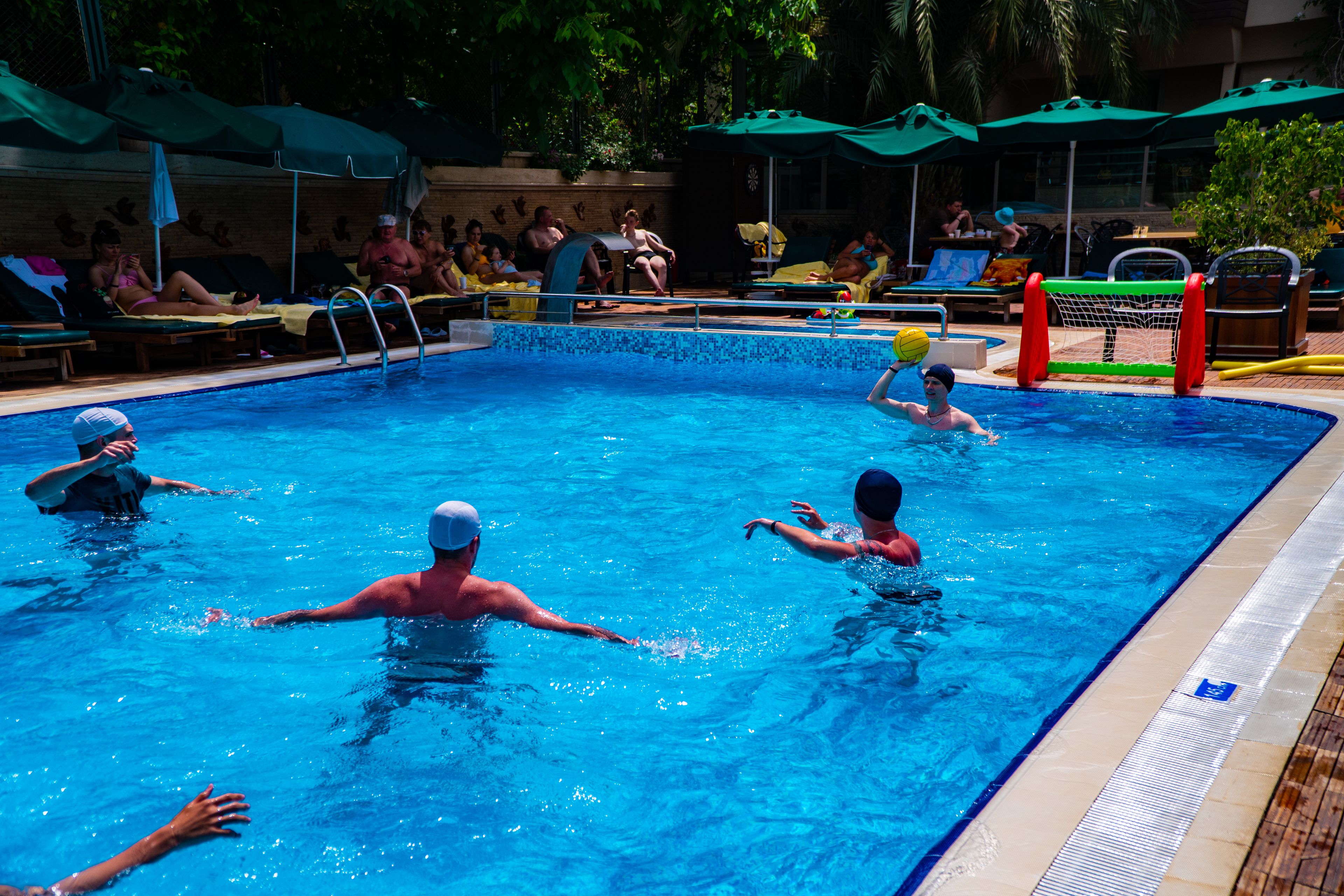 A swimming pool with people in the water, lounge chairs, and umbrellas around the pool area.