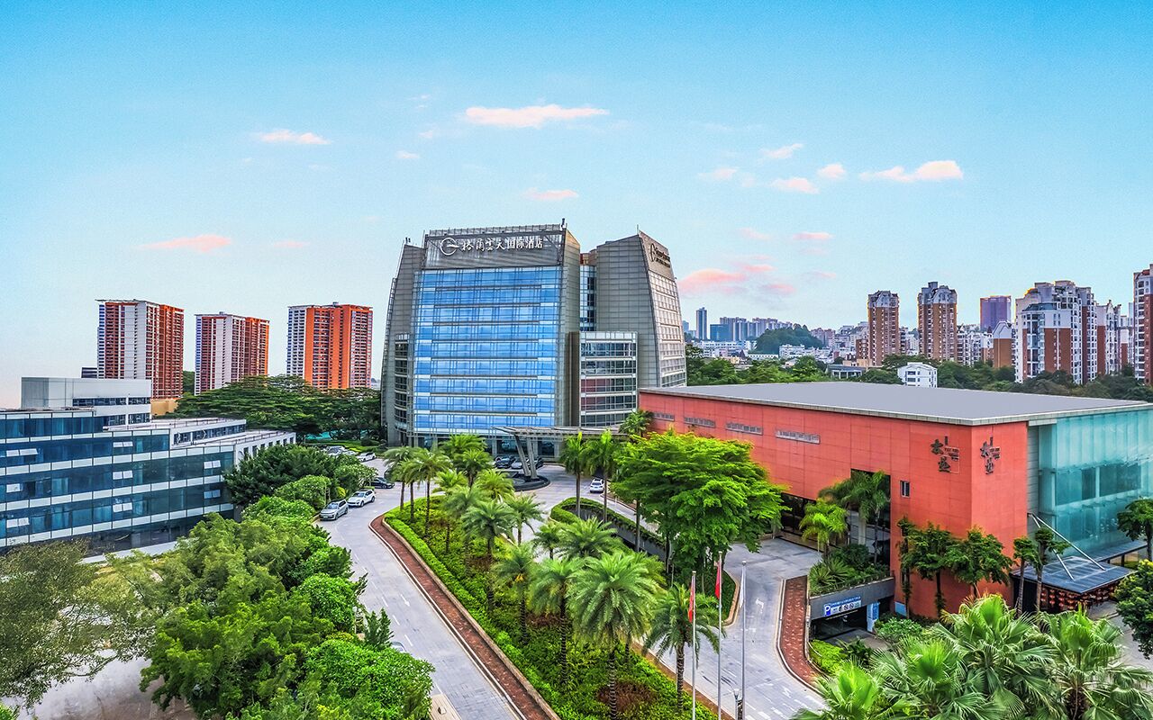 A modern building complex with a prominent glass facade, surrounded by palm trees and other greenery.