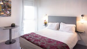 A neatly arranged hotel room with a large bed, a small side table with a plant, a modern chair, and a wall-mounted light fixture. - Nemea Appart’Hotel Toulouse Constellation (Toulouse)