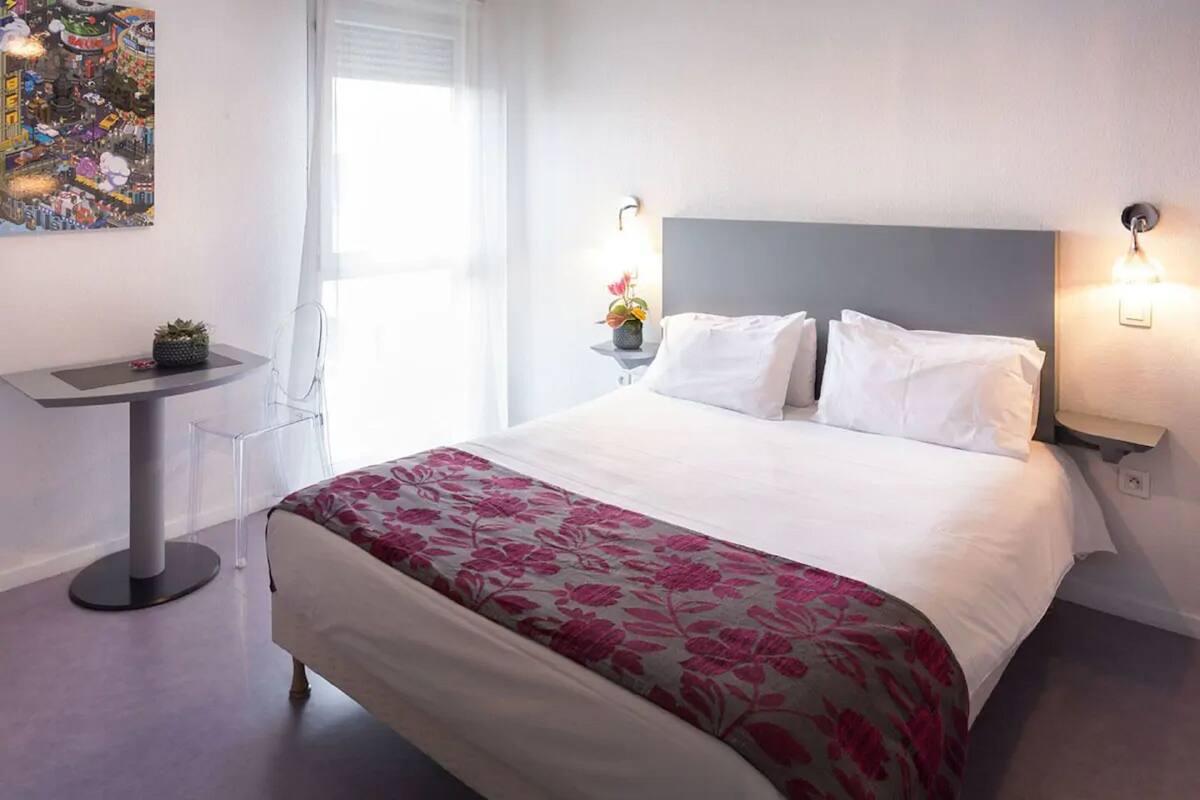 Een netjes ingerichte hotelkamer met een groot bed, een klein bijzettafeltje met een plant, een moderne stoel en een wandlamp.