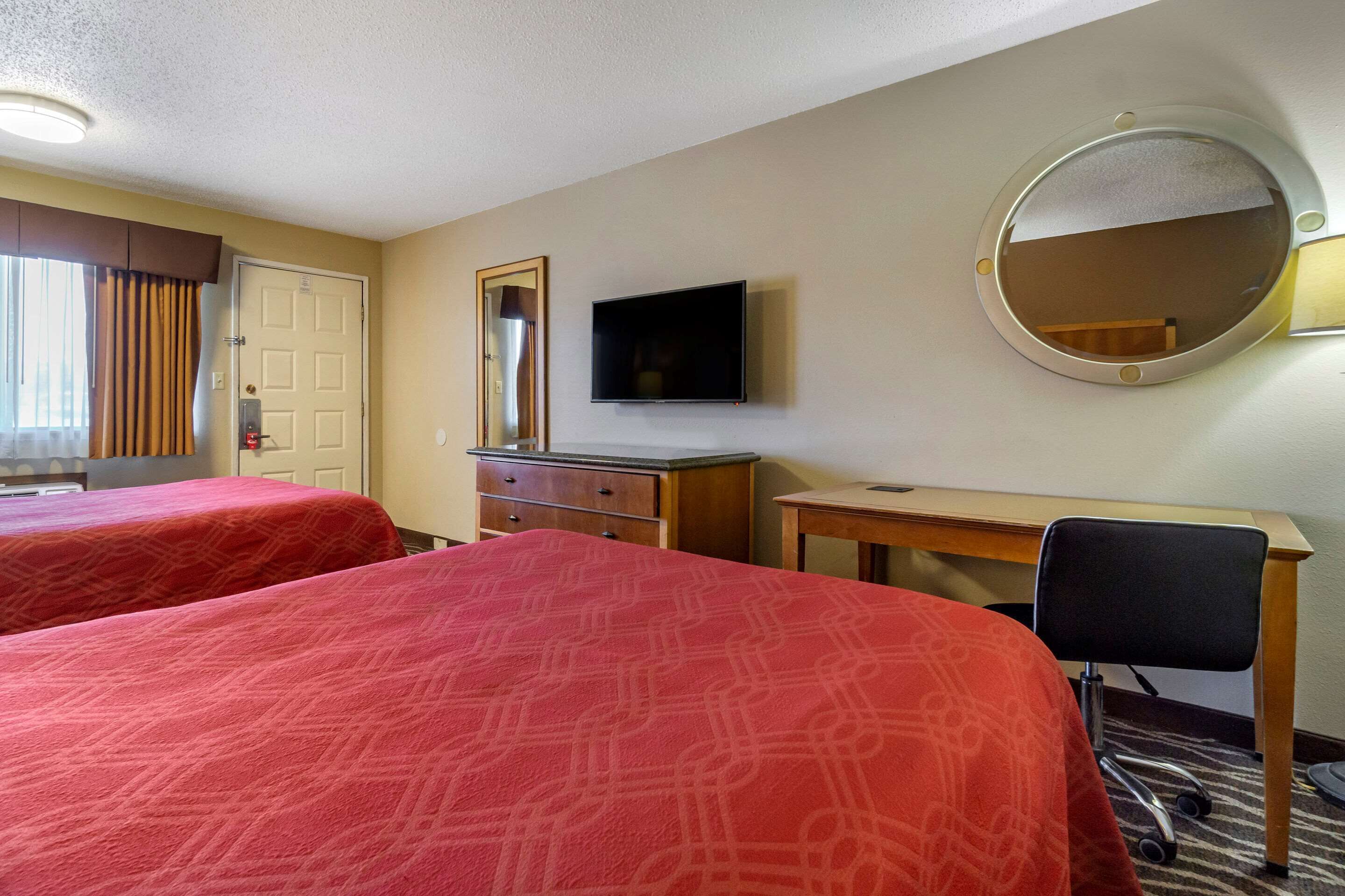 Quarto de hotel com duas camas, uma escrivaninha, uma cadeira, televisão, espelho e porta.