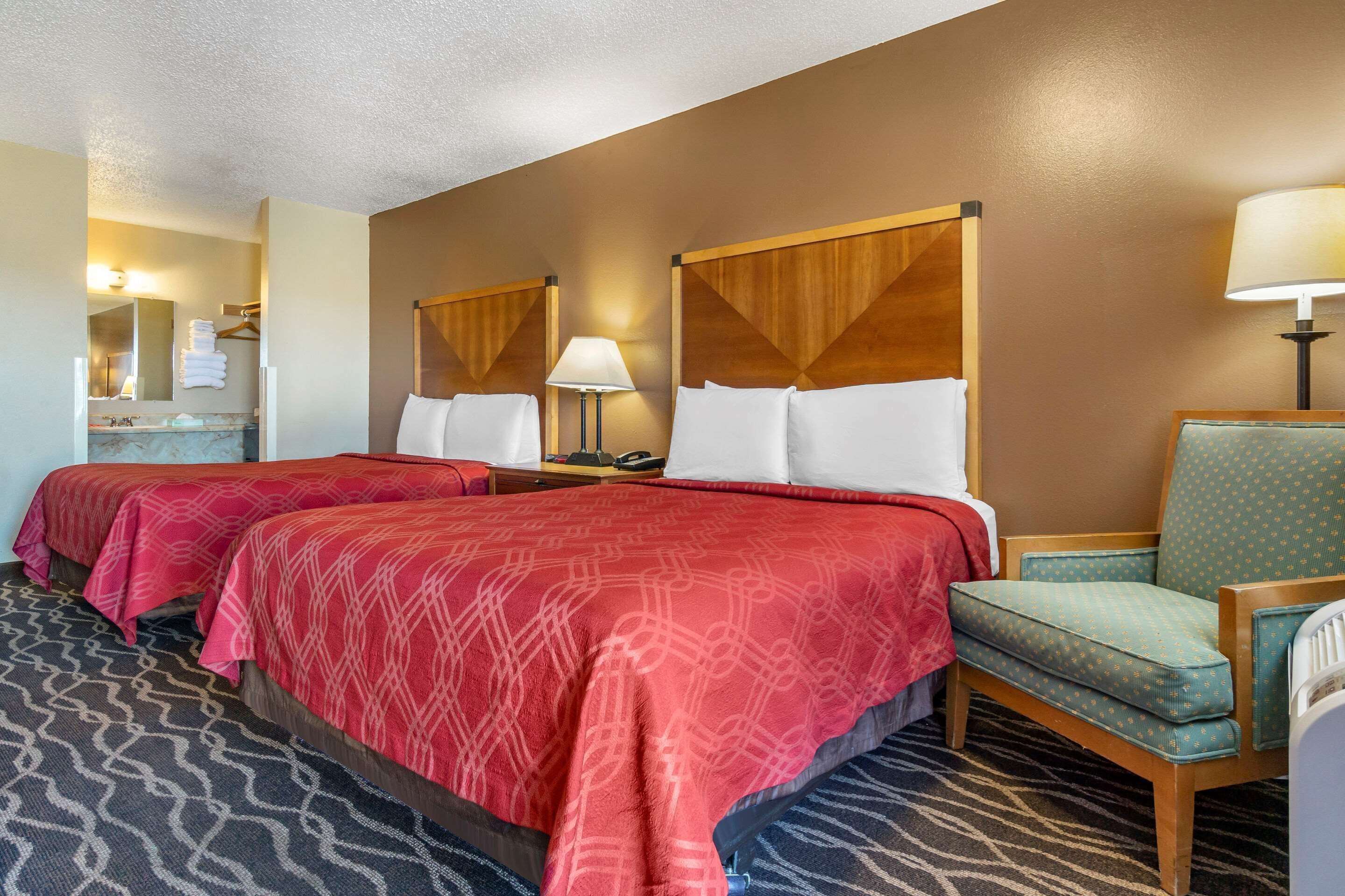 A hotel room with two beds, a sitting area with a chair, a nightstand, a lamp, and a bathroom with a mirror and towel rack.