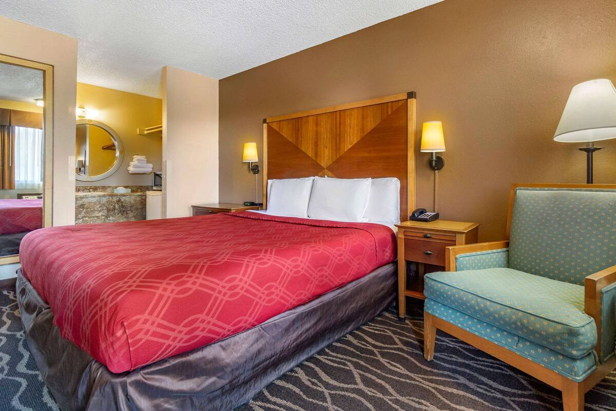 Une chambre d’hôtel avec un grand lit, une tête de lit en bois, deux lampes de chevet, une petite table, une chaise et une salle de bain avec un miroir et un lavabo.