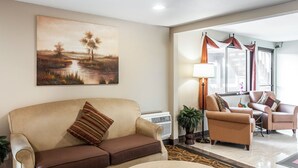 A living room with a beige sofa, a brown armchair, and a round coffee table. There is a painting on the wall, a lamp, and a potted plant.