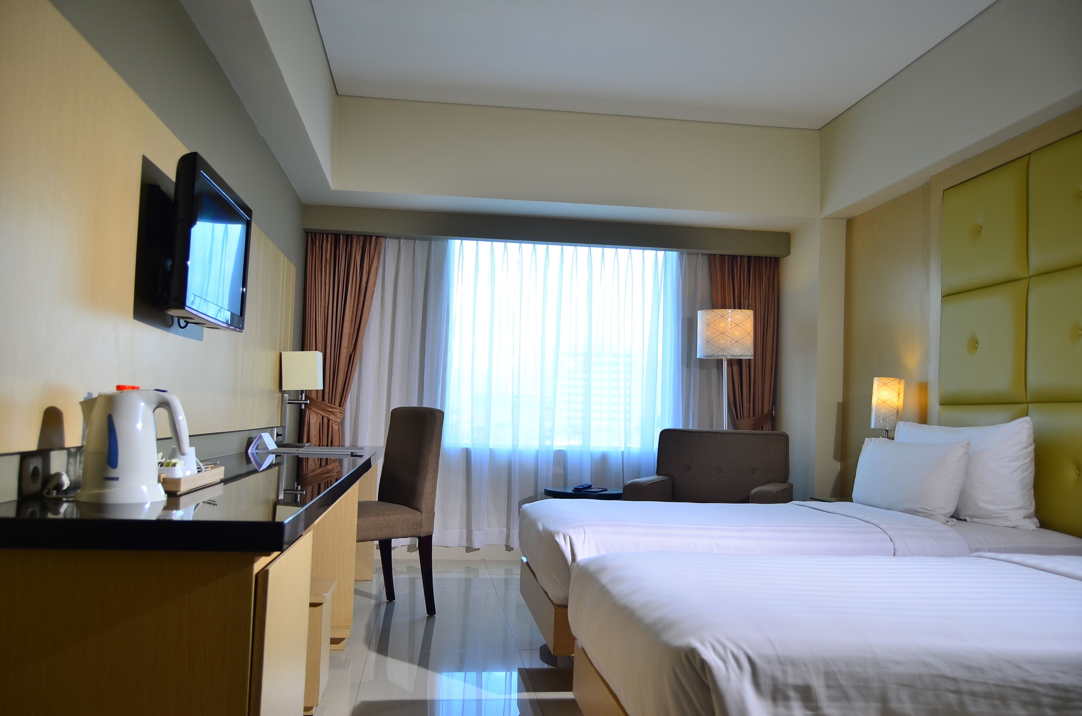 A hotel room with a bed, a desk with a chair, a television mounted on the wall, and a window with curtains.