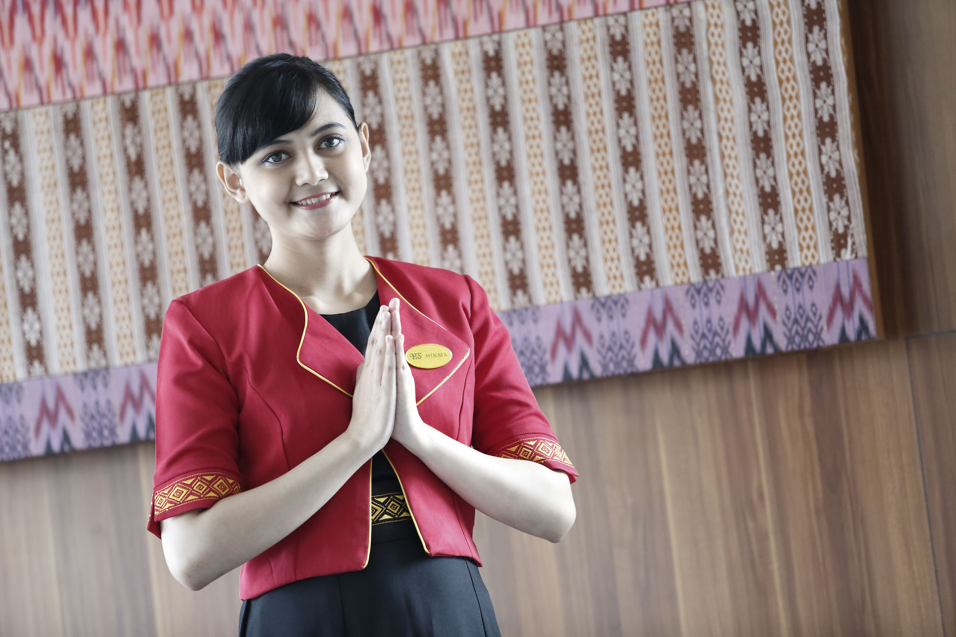 A person in a red uniform with a name tag, standing in front of a patterned wall.