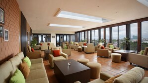 A spacious lounge area with multiple sofas and armchairs arranged around a central coffee table, large windows offering a city view, and a skylight above. - Hotel Santika Makassar (Makassar)