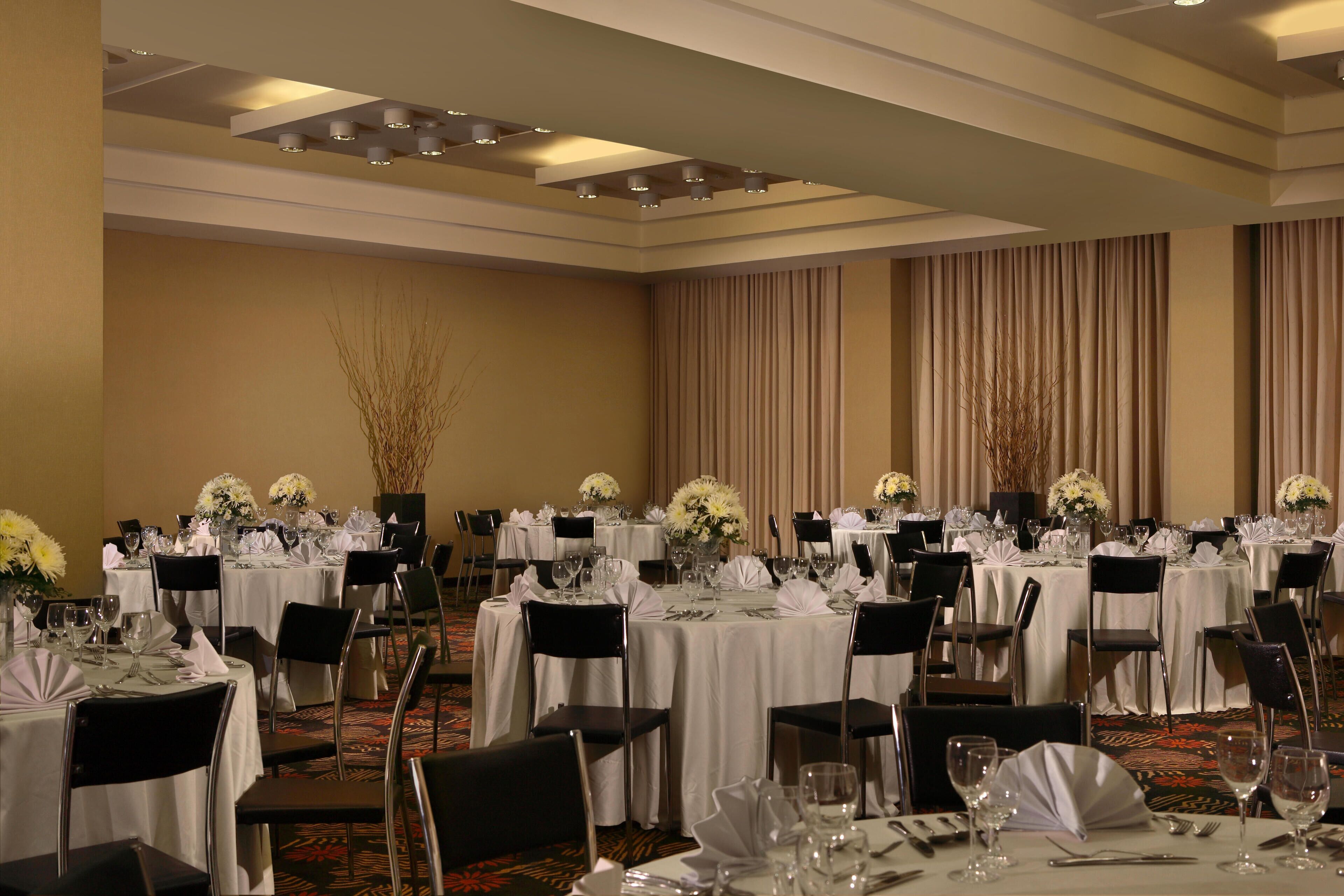 A banquet hall with round tables set for dining, each with white tablecloths, black chairs, and floral centerpieces.