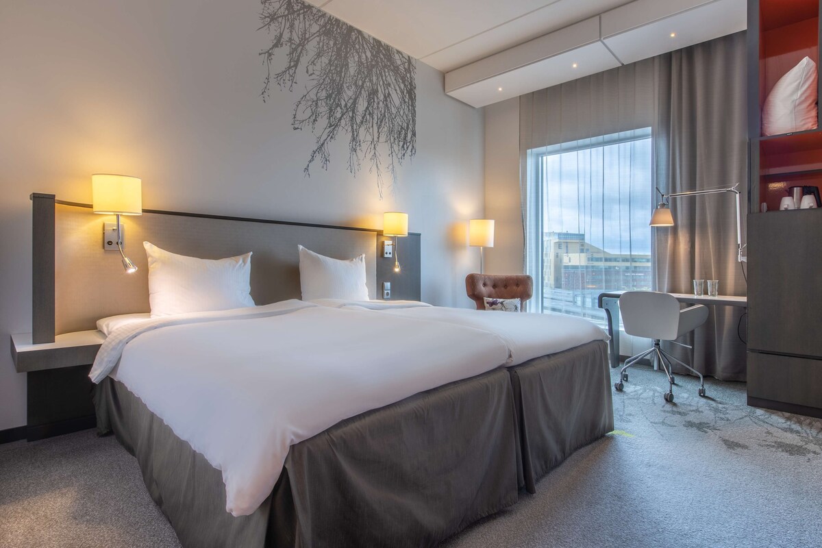 Une chambre d’hôtel avec un grand lit, deux lampes de chevet, un bureau avec une chaise et une vue sur la ville par une grande fenêtre.