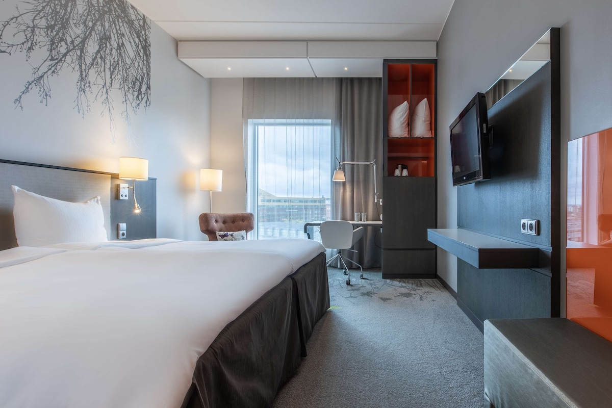 Une chambre d’hôtel avec un grand lit, un bureau, une chaise, une télévision et une fenêtre donnant sur un beau paysage.