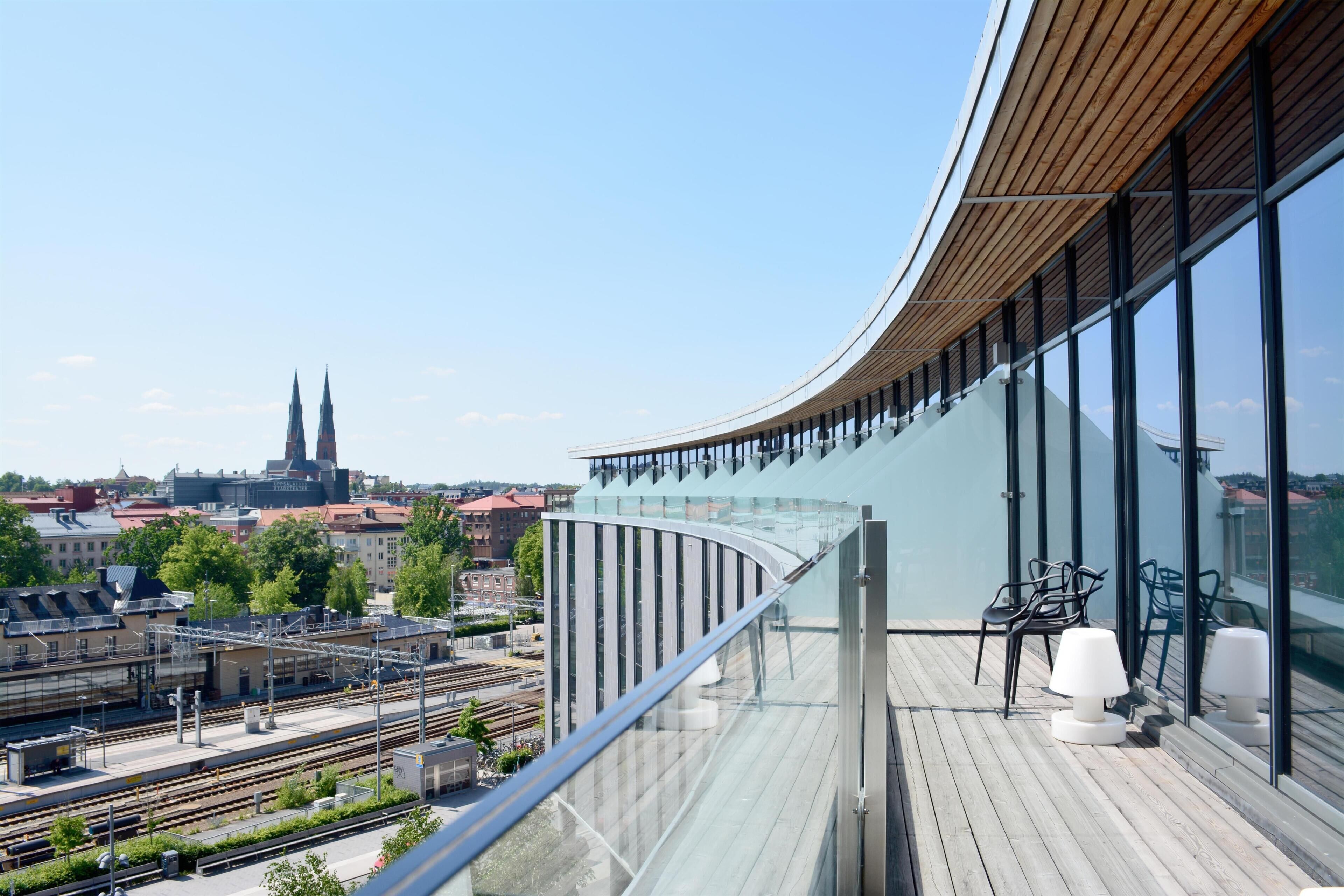 A modern building with a curved glass facade, a balcony with seating, and a view of a train station and cityscape.