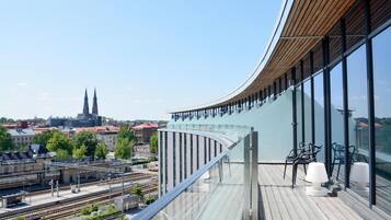 A modern building with a curved glass facade, a balcony with seating, and a view of a train station and cityscape.