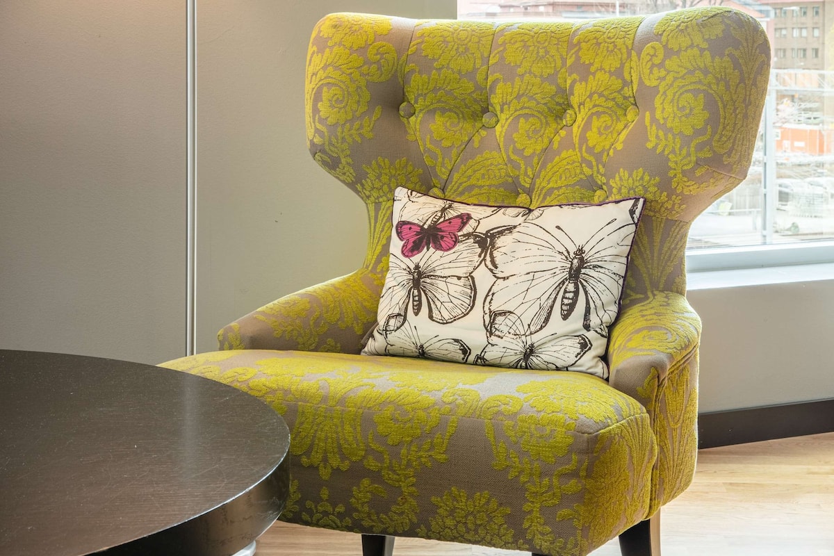 A yellow armchair with a floral pattern and a butterfly pillow, positioned next to a round table and a floor lamp, with a cityscape view through a window.