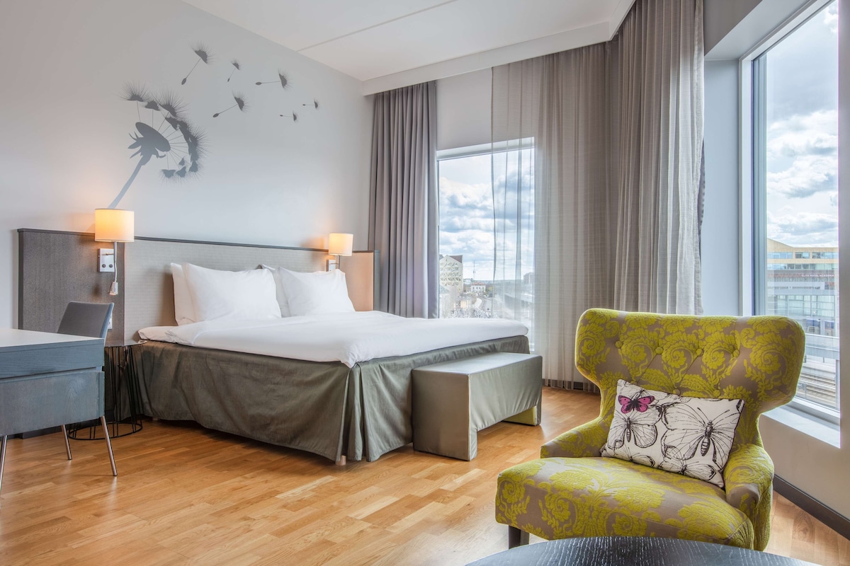 Une chambre d’hôtel avec un lit, des lampes de chevet, une chaise, un bureau et une grande fenêtre.