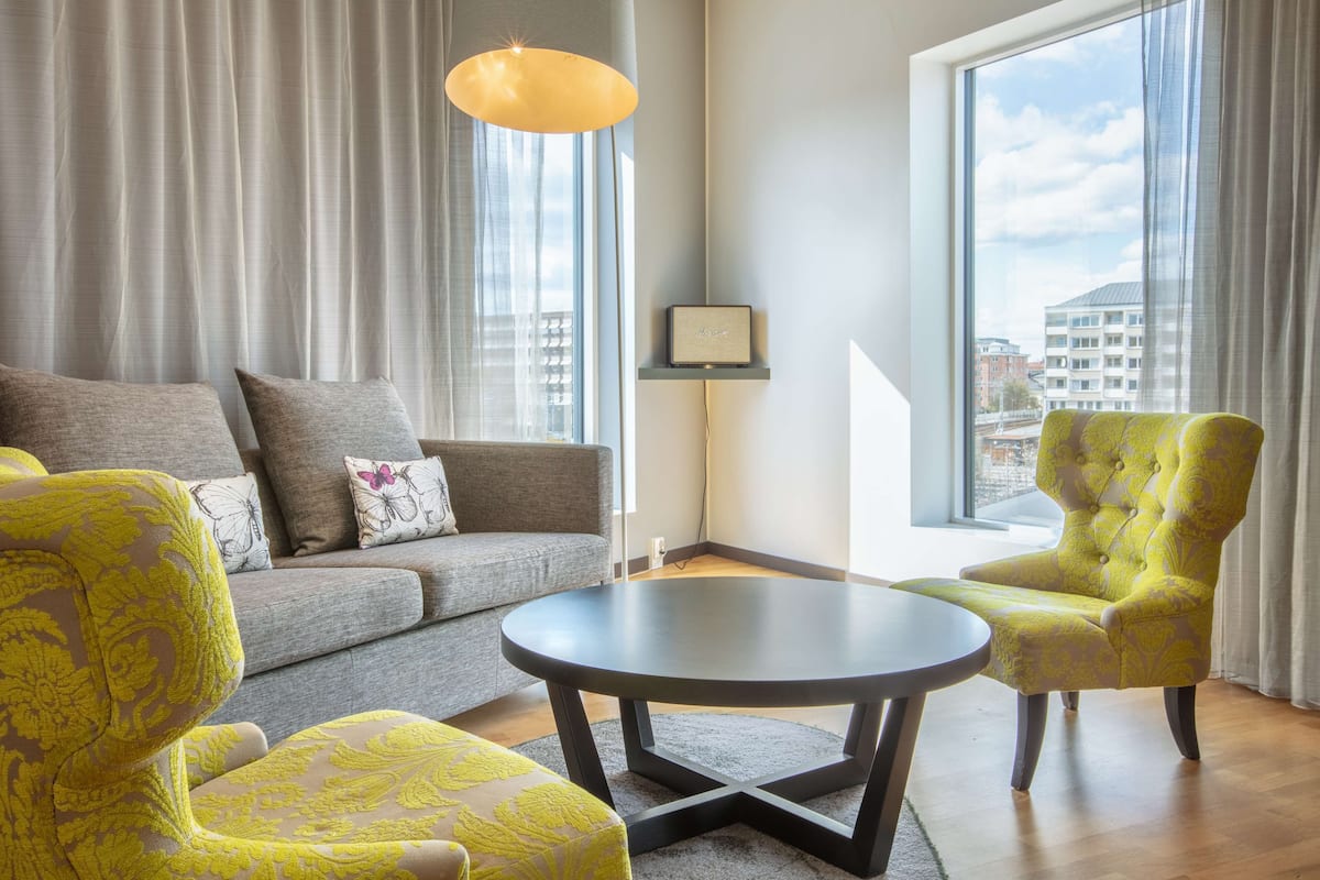 A modern living room with a grey sofa, yellow patterned armchairs, a round coffee table, and a large window with a city view.