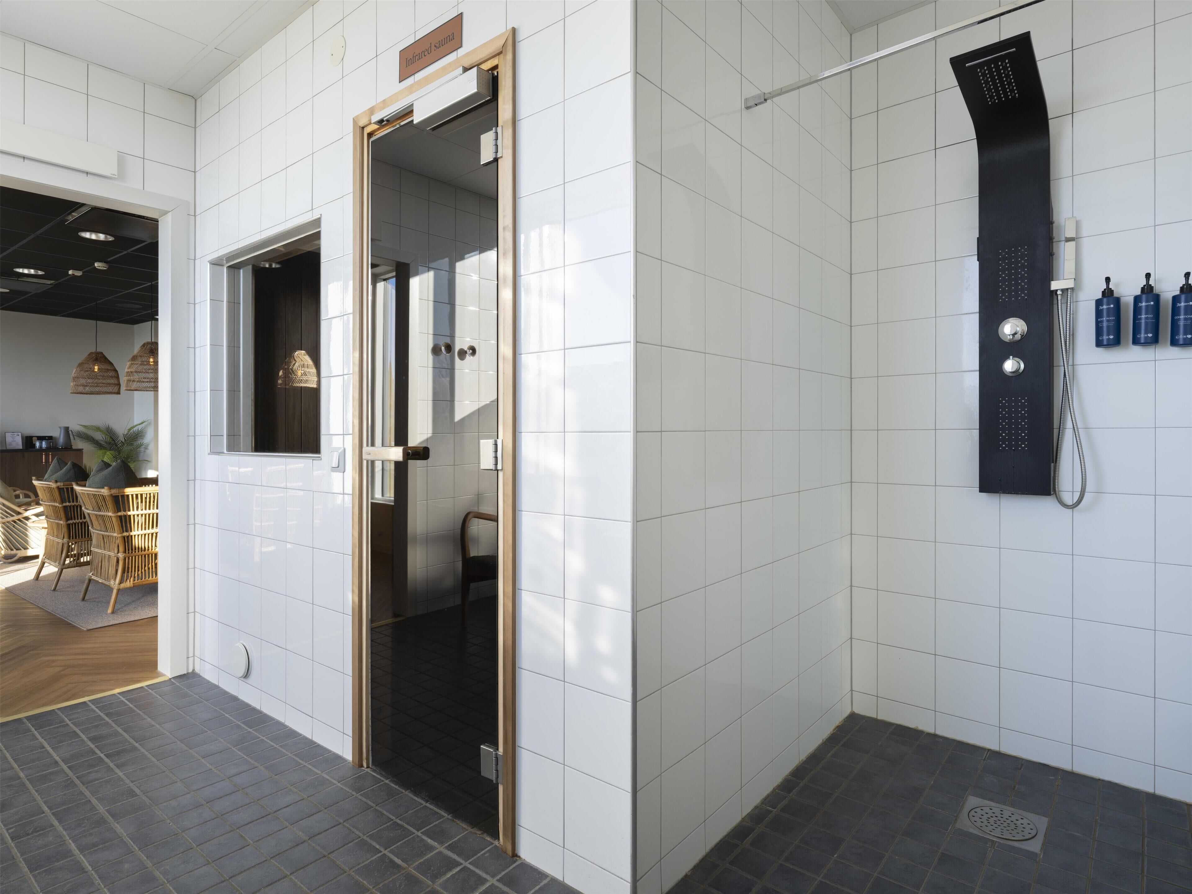 A modern bathroom with white tiled walls, a black shower panel, and a visible infrared sauna sign.