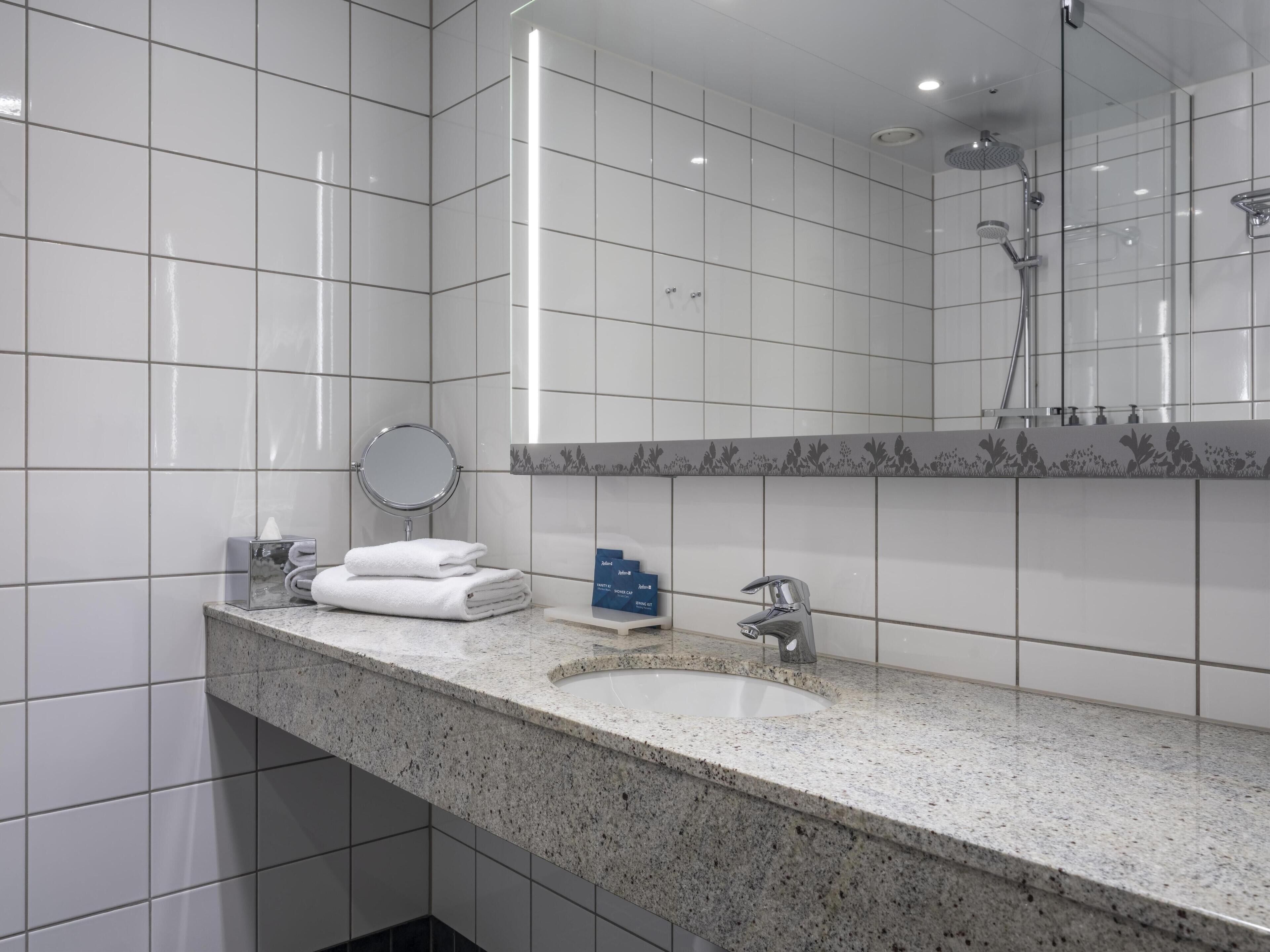 Ein modernes Badezimmer mit weißen Fliesen, einer Granit-Arbeitsplatte, einem runden Spiegel, einer Handbrause und einem Waschbecken mit Armatur.