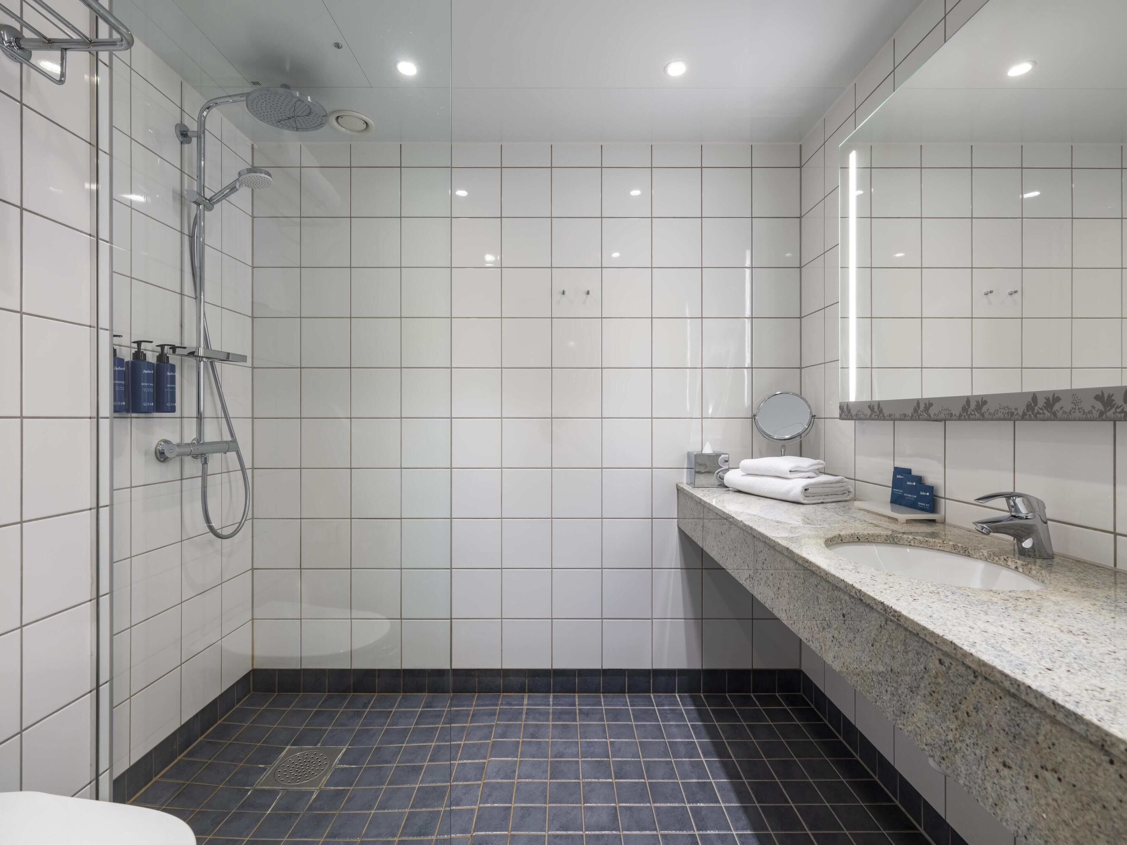 Une salle de bain moderne avec des carreaux blancs, une douche, un lavabo doté d’un plan de travail en marbre et un miroir.