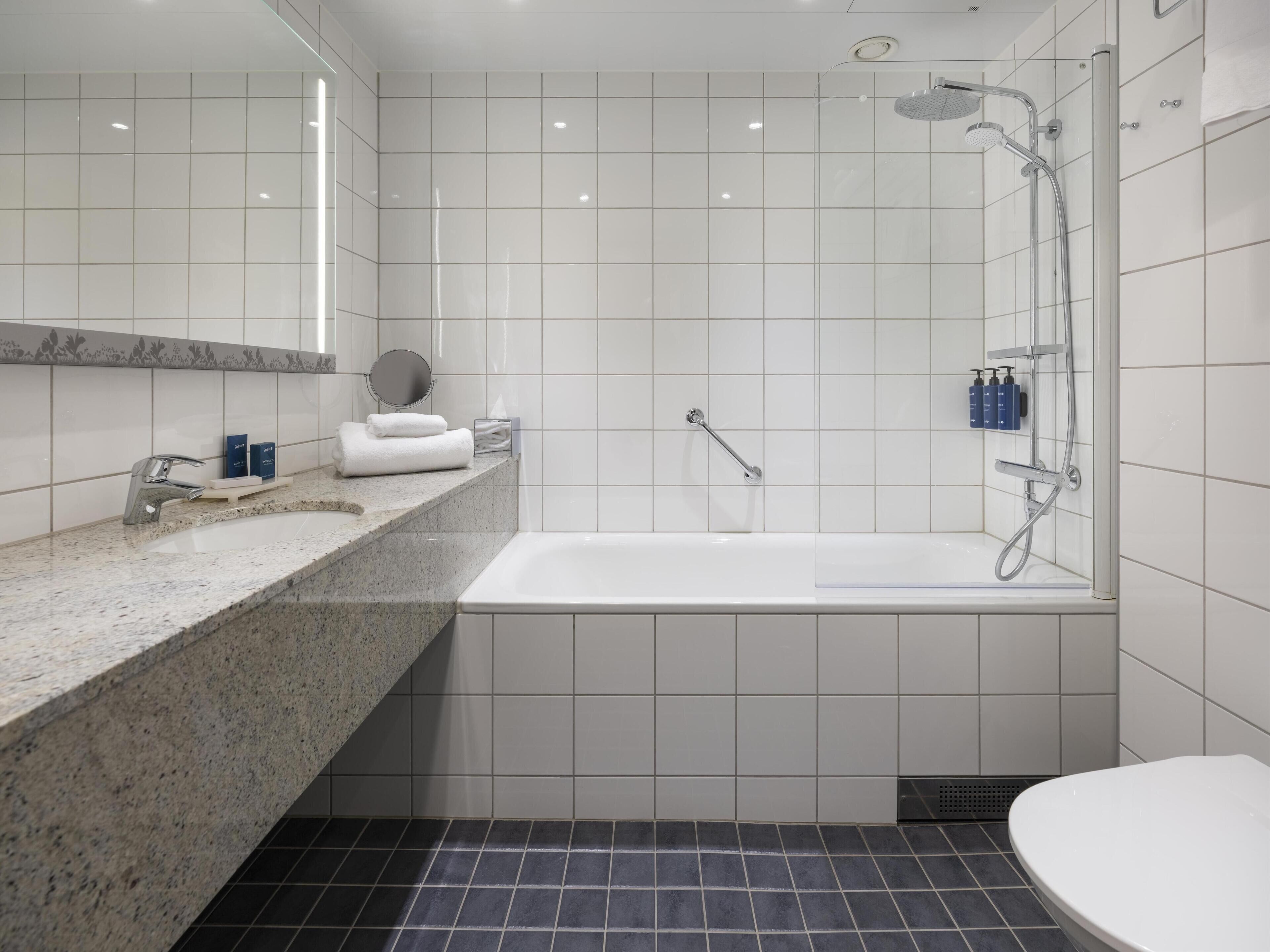 Un baño moderno con encimera de granito, azulejos blancos, bañera, ducha y retrete.