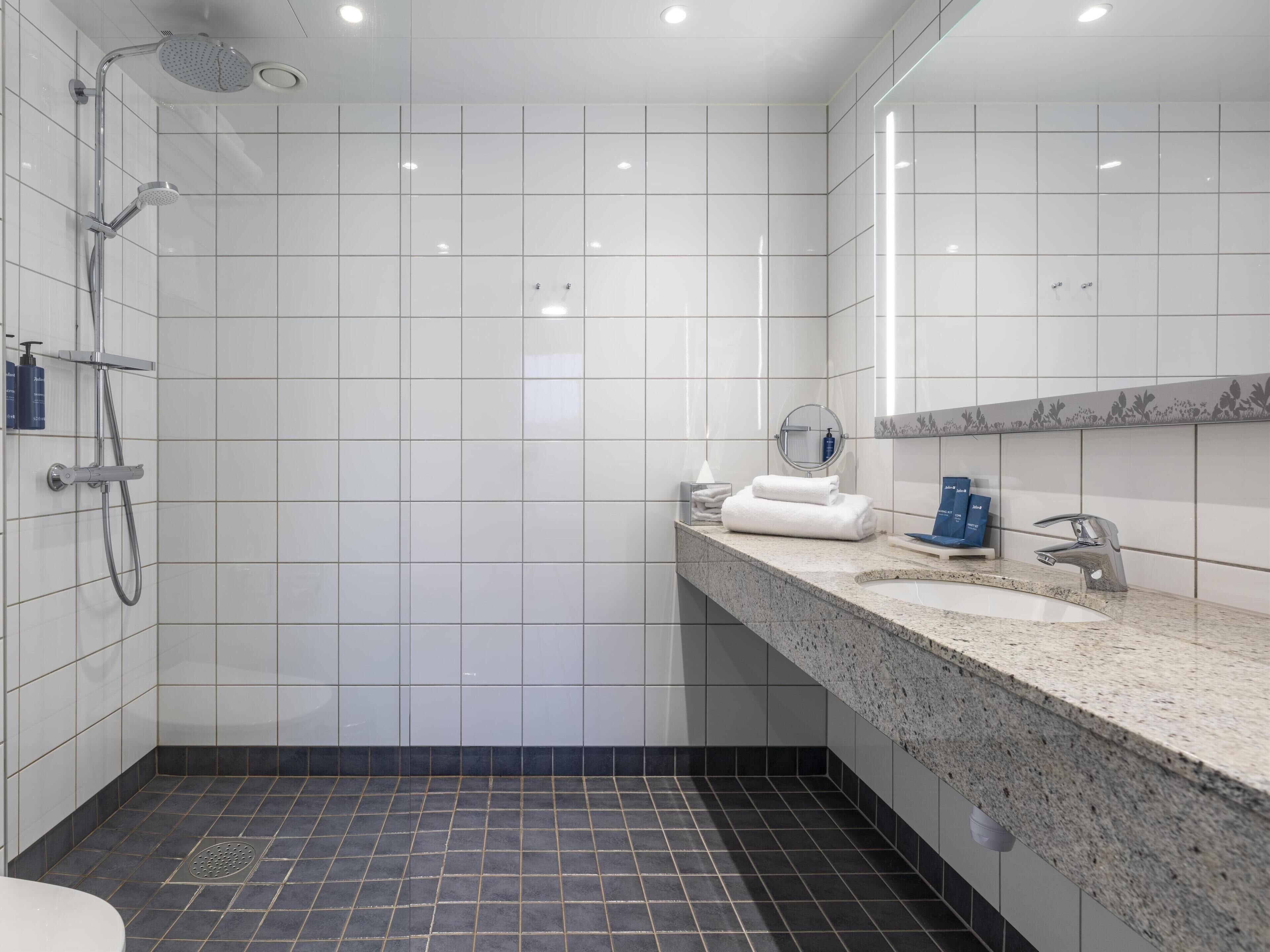 Une salle de bain moderne avec des carreaux blancs, une douche, un lavabo doté d’un plan de travail en granit et un miroir.