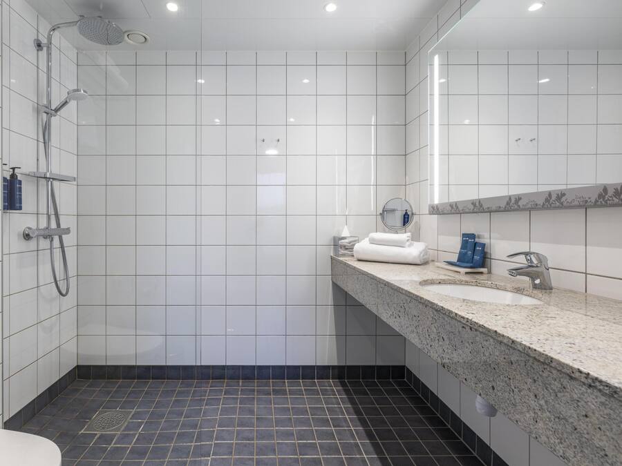 Une salle de bain moderne avec des carreaux blancs, une douche, un lavabo doté d’un plan de travail en granit et un miroir.