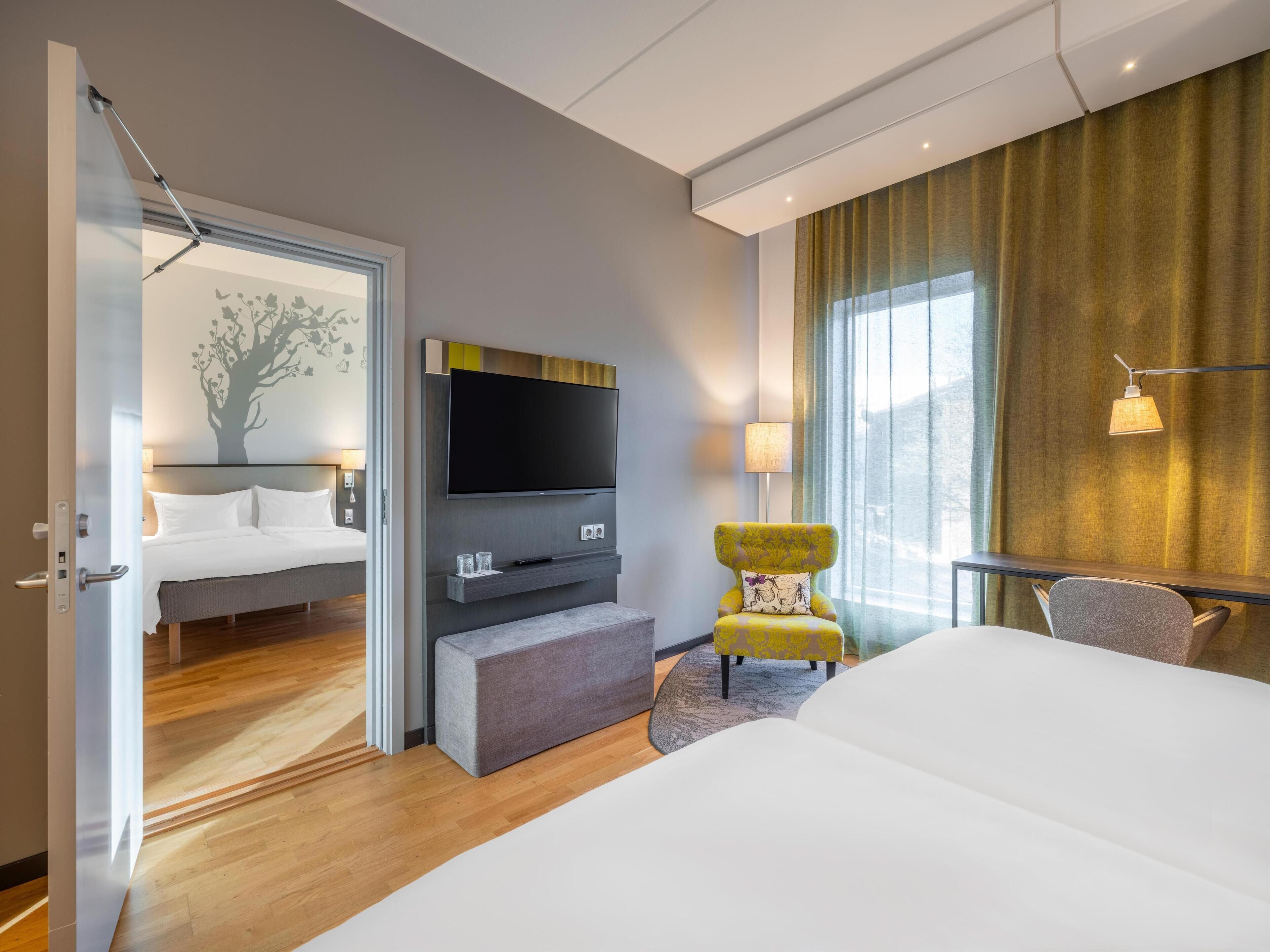 A modern hotel room with a large bed, a flat-screen TV mounted on the wall, a small seating area with a chair, and a large window with curtains.
