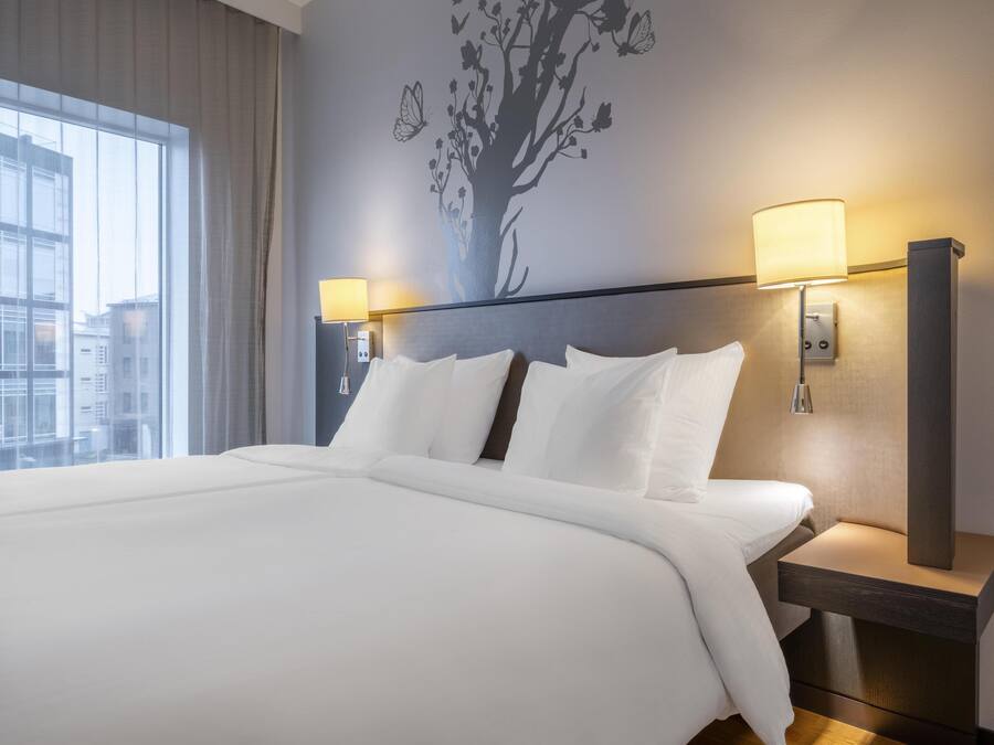 Una habitación de hotel con una cama grande, dos lámparas de noche, un mural de un árbol y una ventana con cortinas.