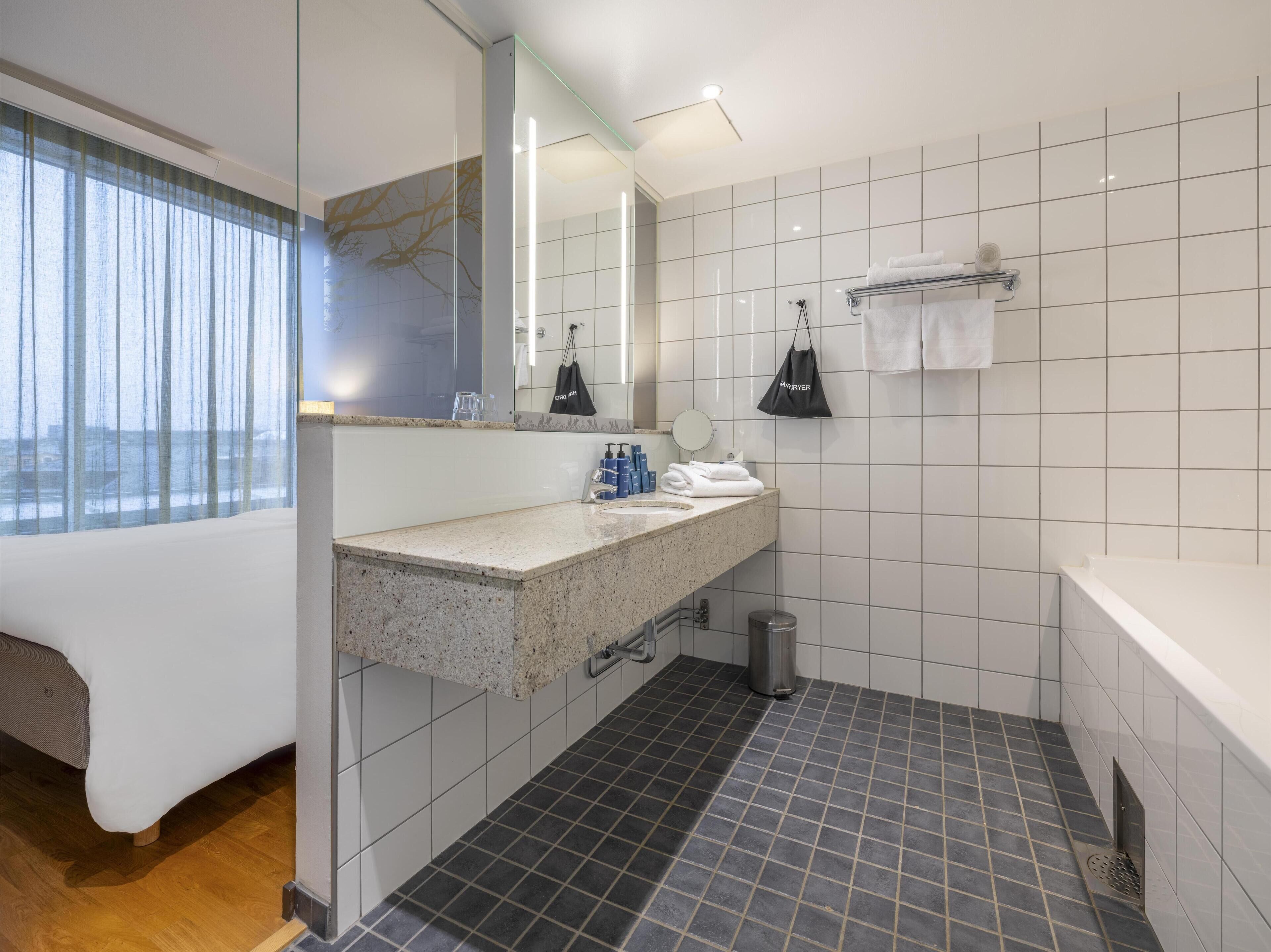 A modern bathroom with a granite countertop, a large mirror, and a white sink. There is a black bag hanging on the wall and a towel rack with towels.