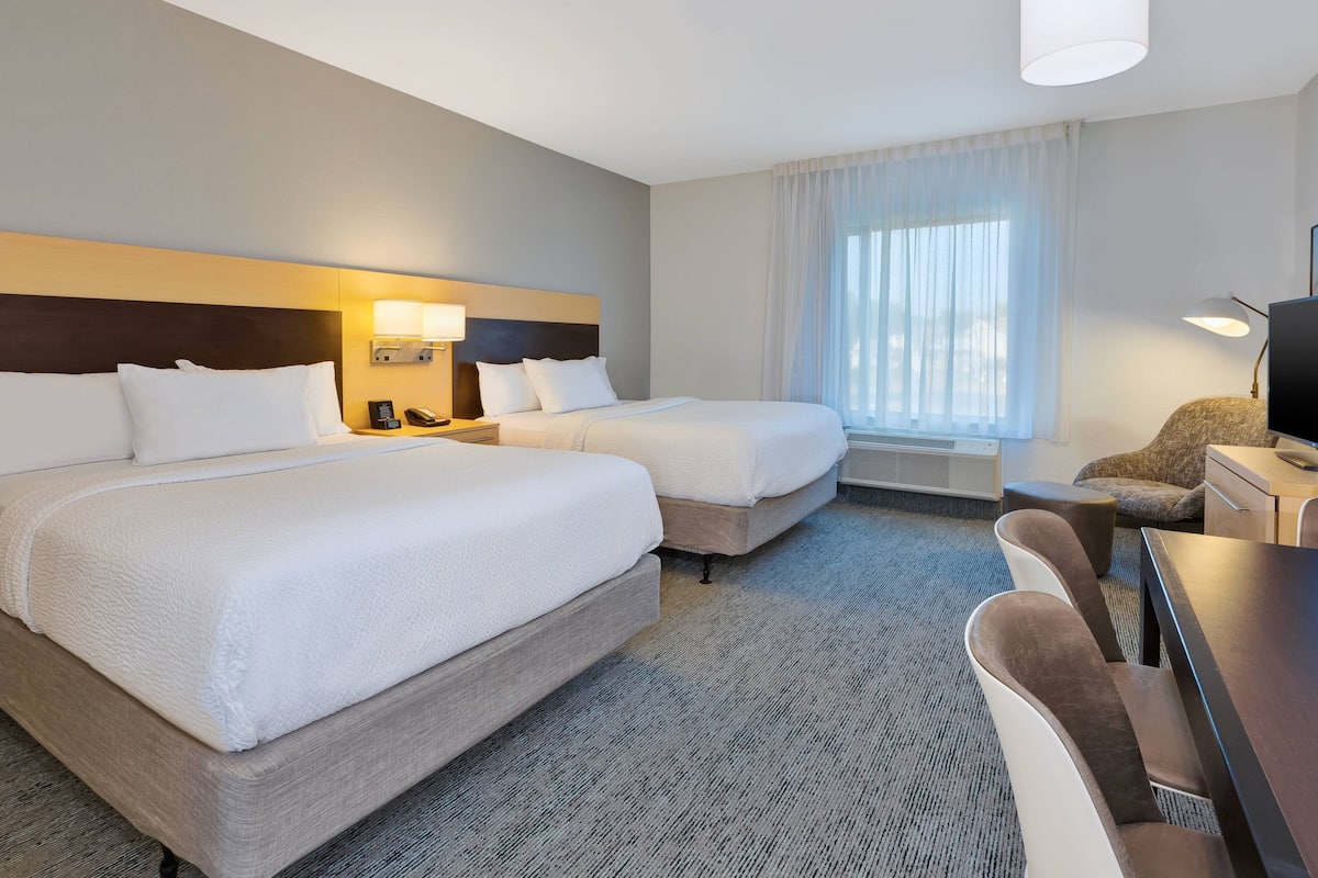 A hotel room with two beds, a desk, a chair, a lamp, and a large window with curtains.