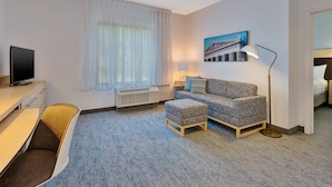 Habitación de hotel con escritorio, sofá, otoman y lámpara de pie, además de un cuadro en la pared.