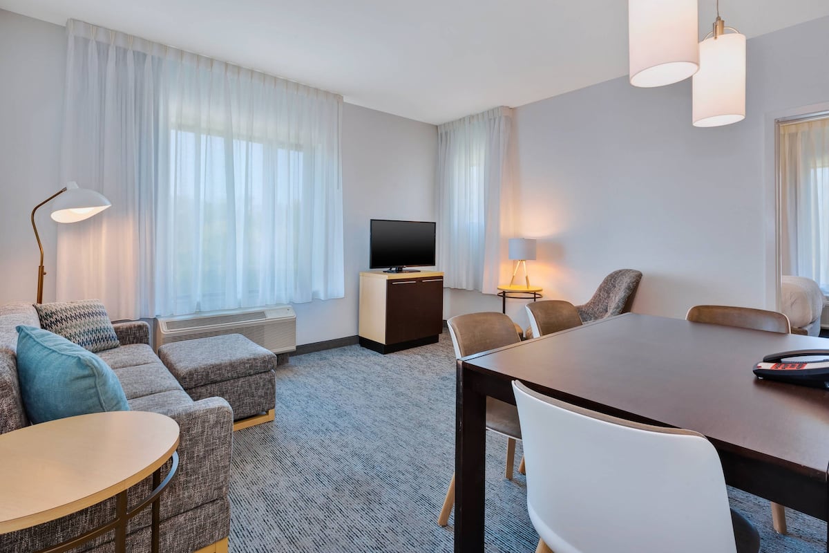 A hotel room with a sofa, a small round table, a larger dining table, a TV, and a lamp.