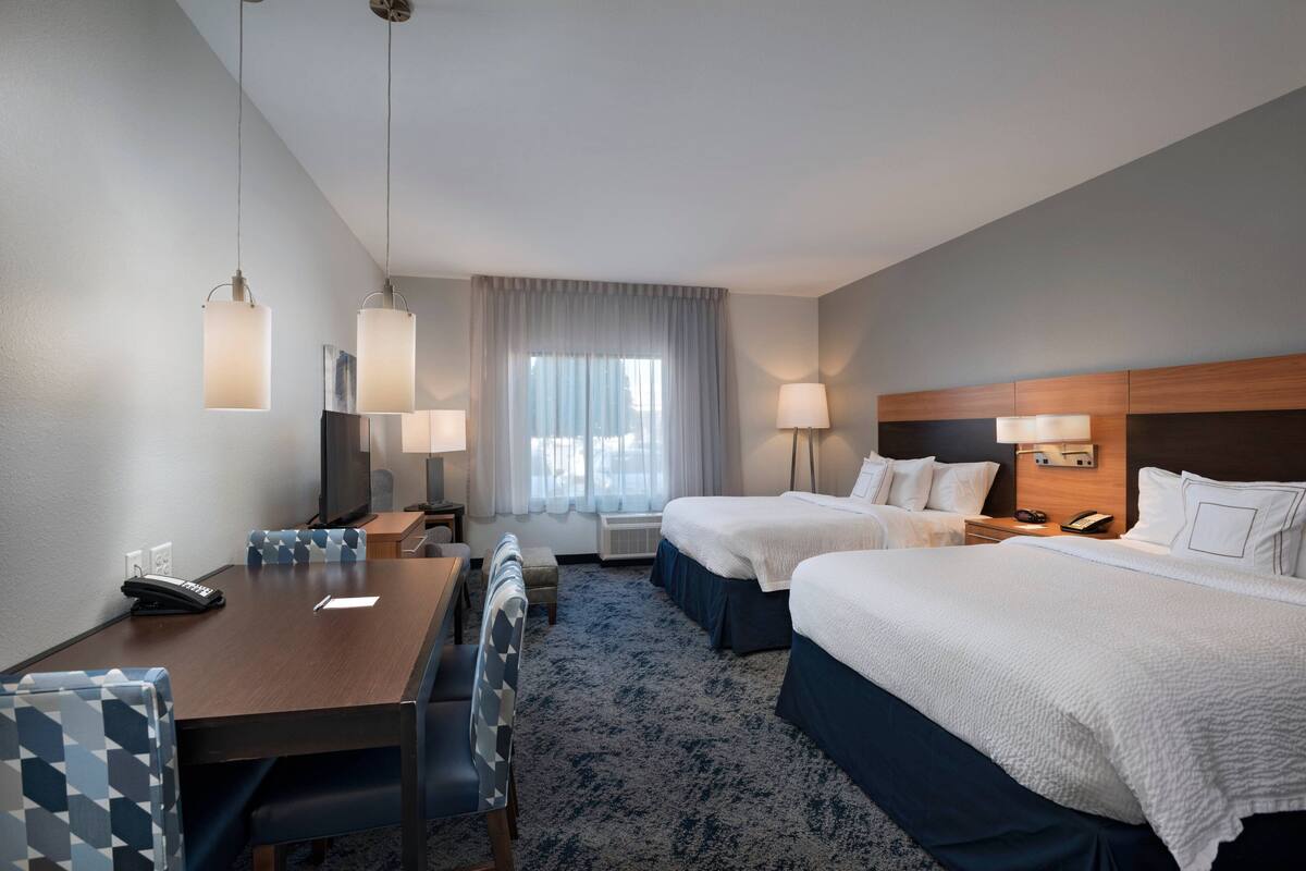A hotel room with two beds, a desk, chairs, a TV, and a window with curtains.