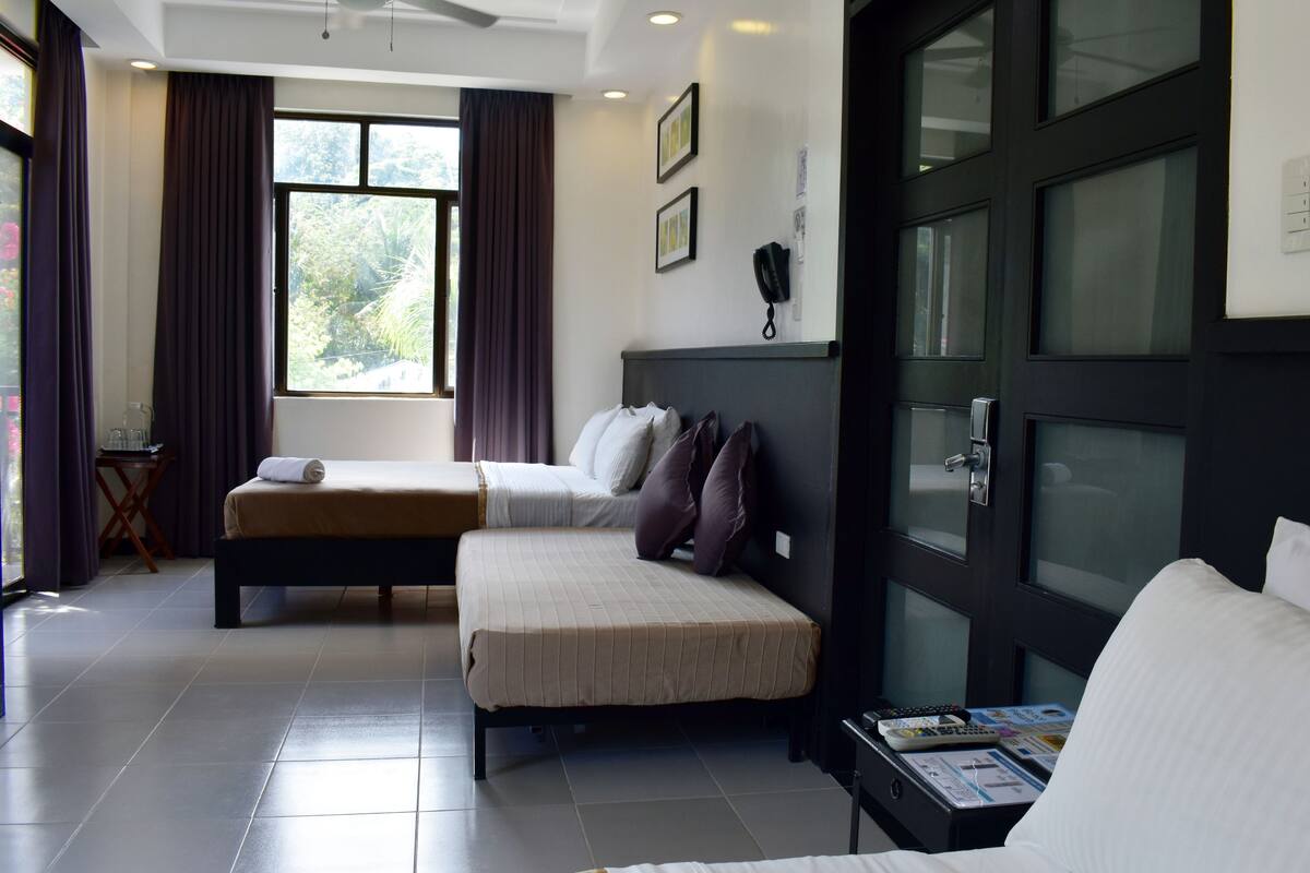 A hotel room with a bed, a sofa, a small table with a remote control, and a window with curtains.