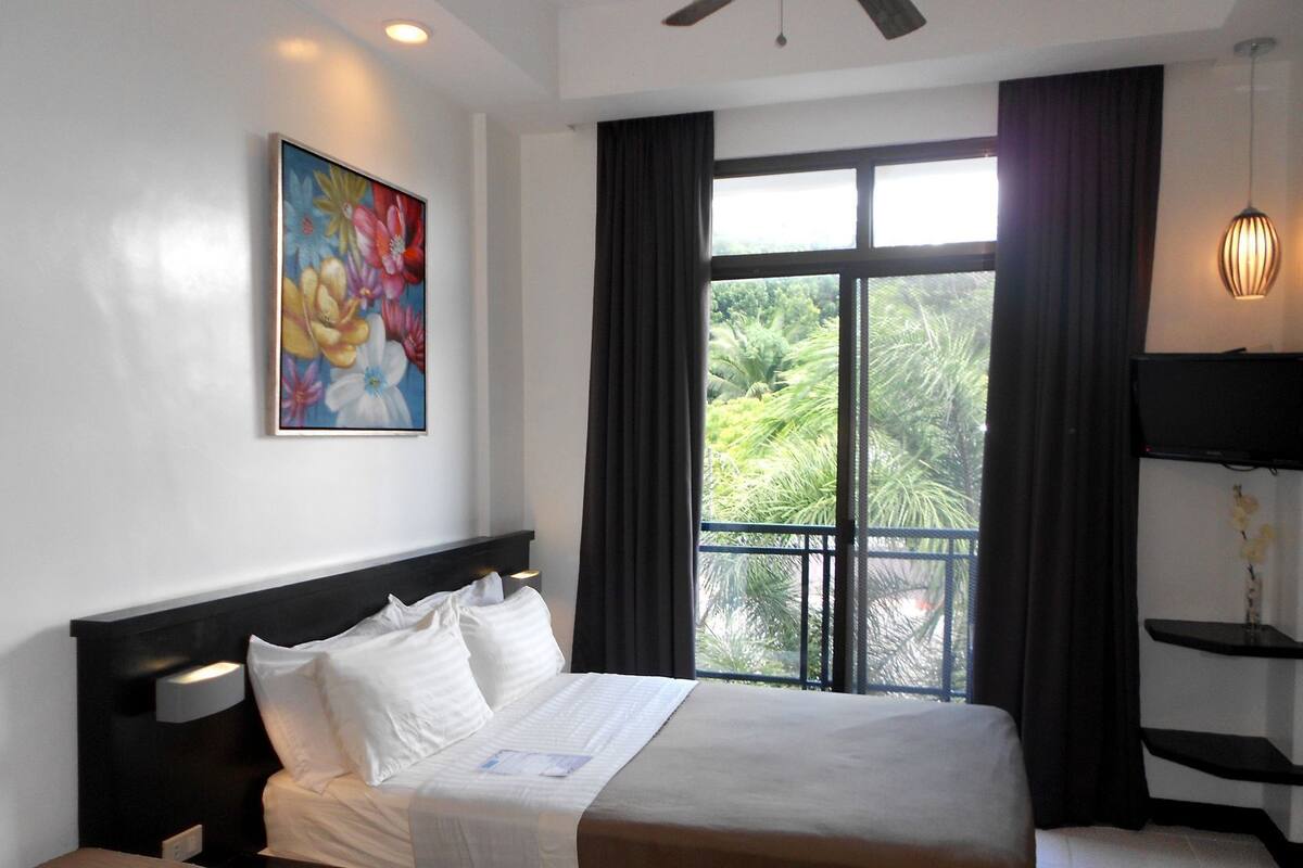 A hotel room with a bed, a painting, a television, and a window with curtains.