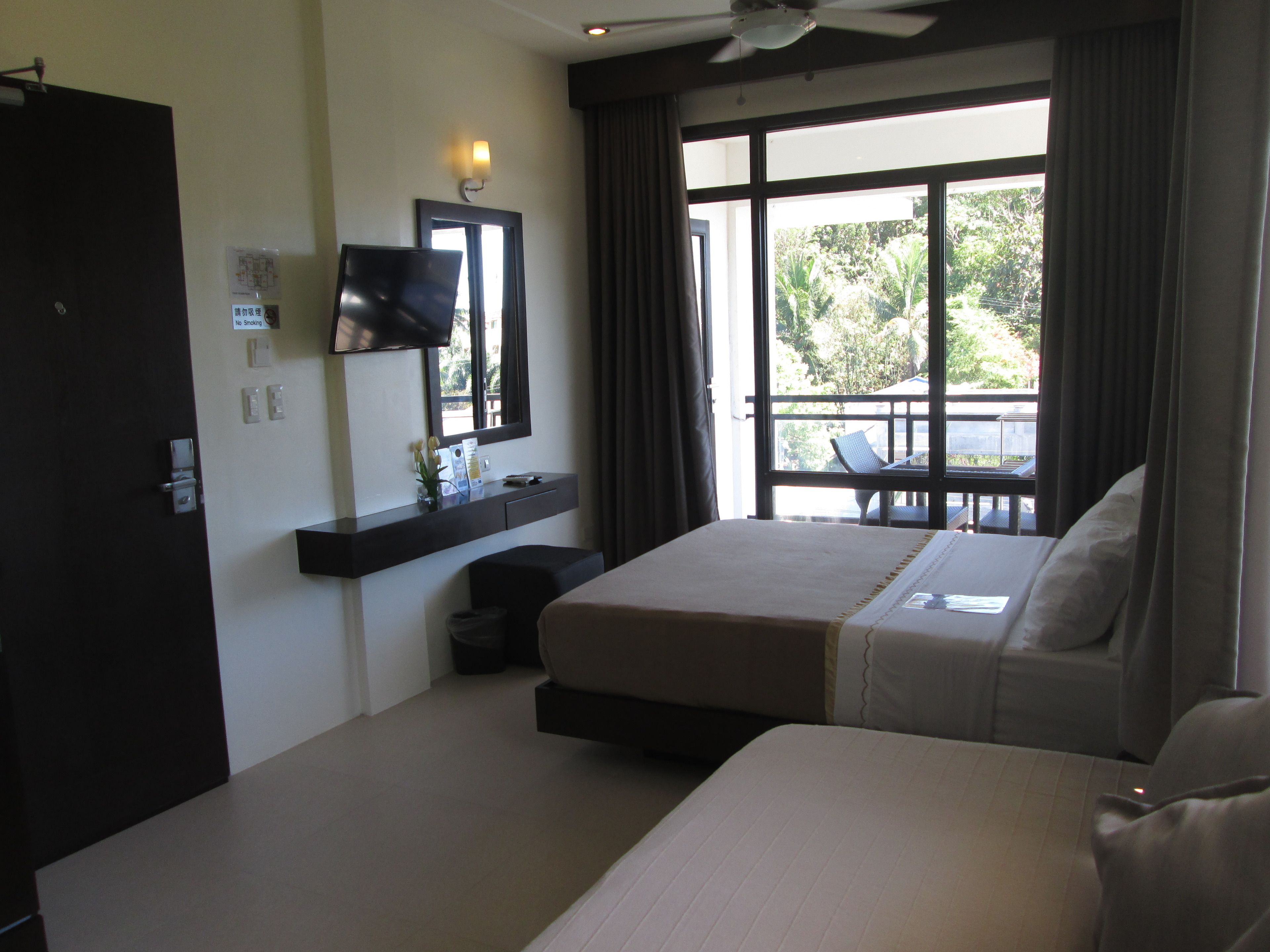 Une chambre d’hôtel avec un lit, un téléviseur fixé au mur, une fenêtre avec des rideaux et un balcon avec des chaises.