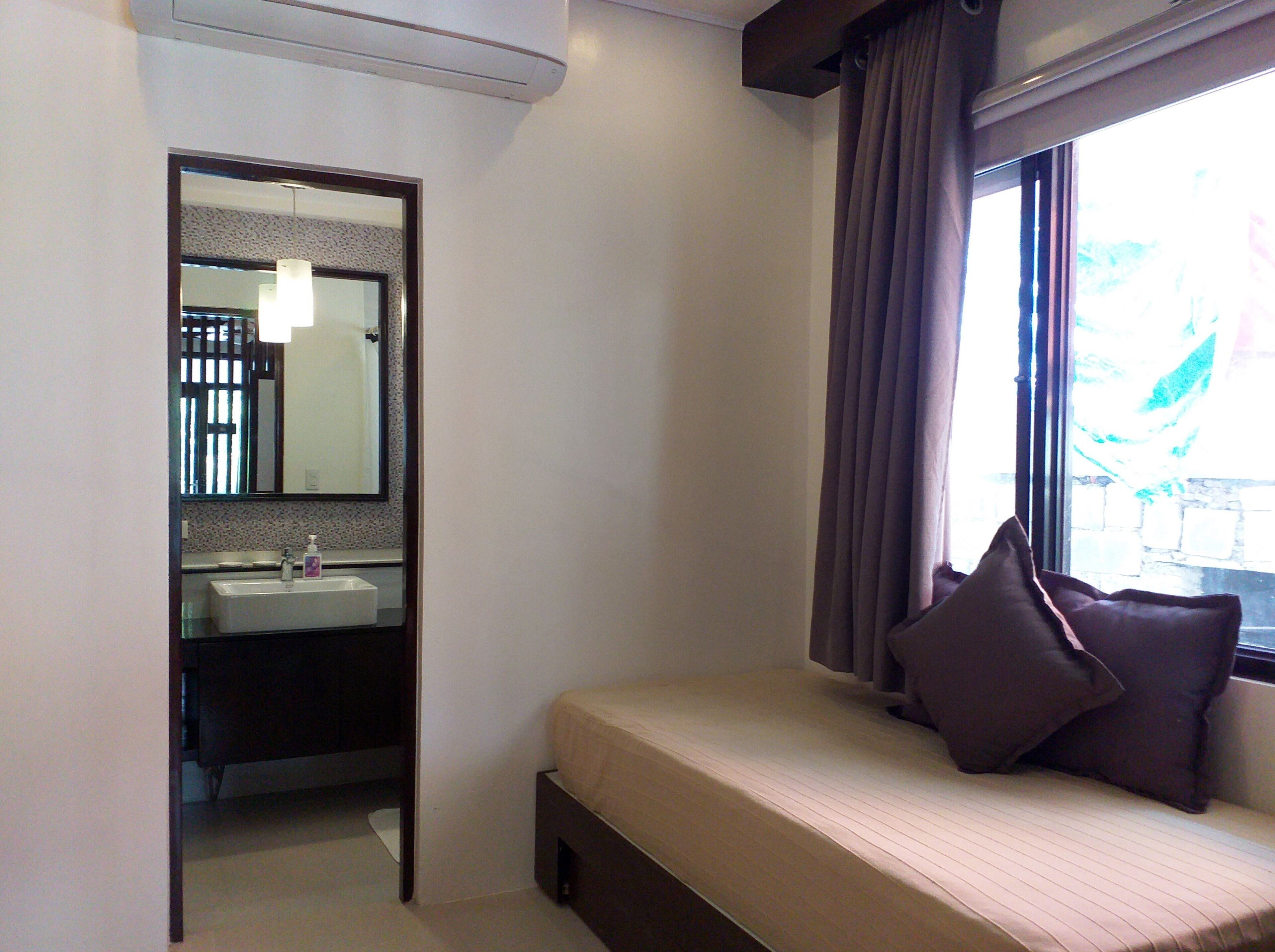 Habitación de hotel con cama, ventana con cortinas, baño con lavamanos y espejo, y una puerta que da a otra habitación.