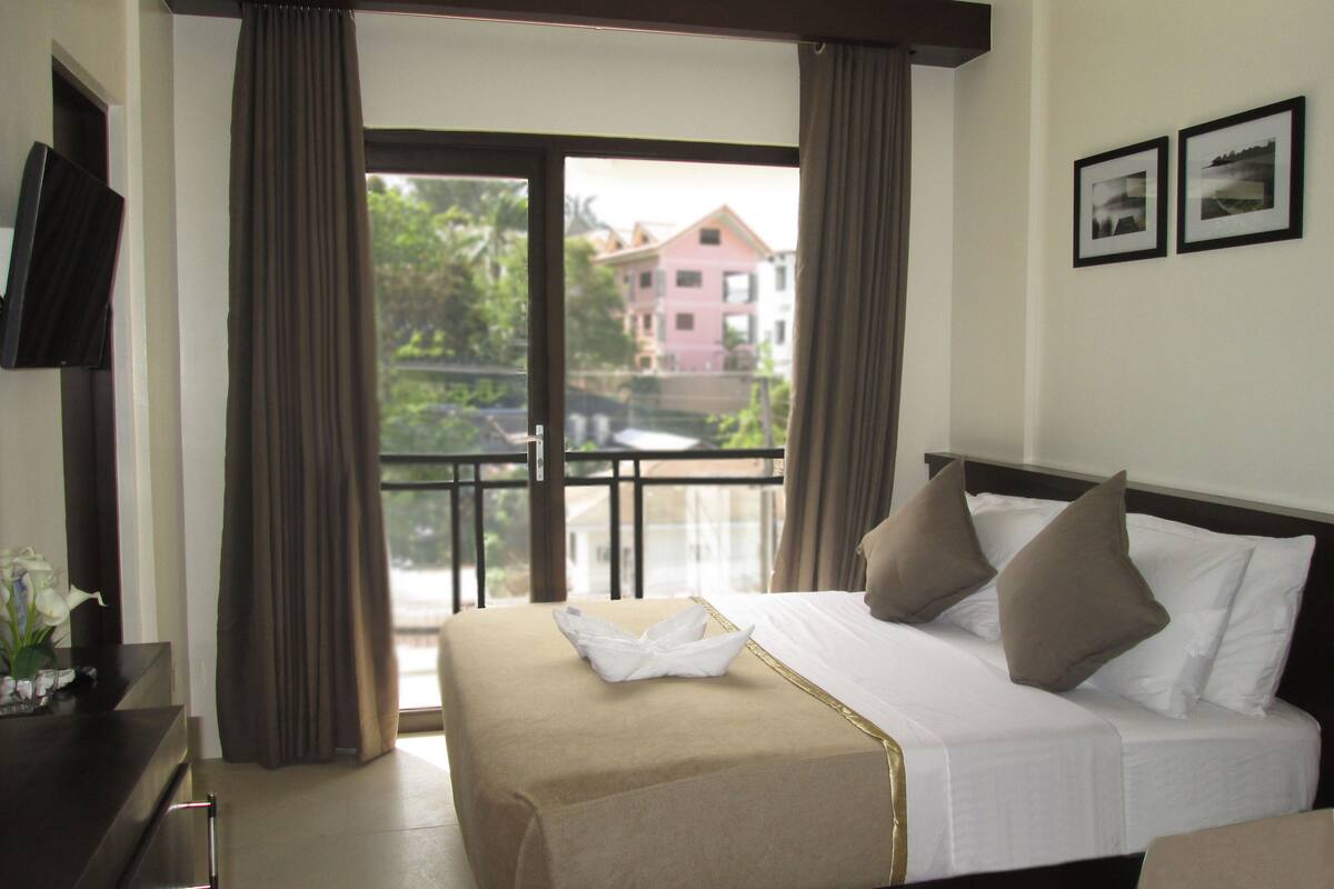 A hotel room with a bed, a TV, a balcony with curtains, and a view of buildings outside.